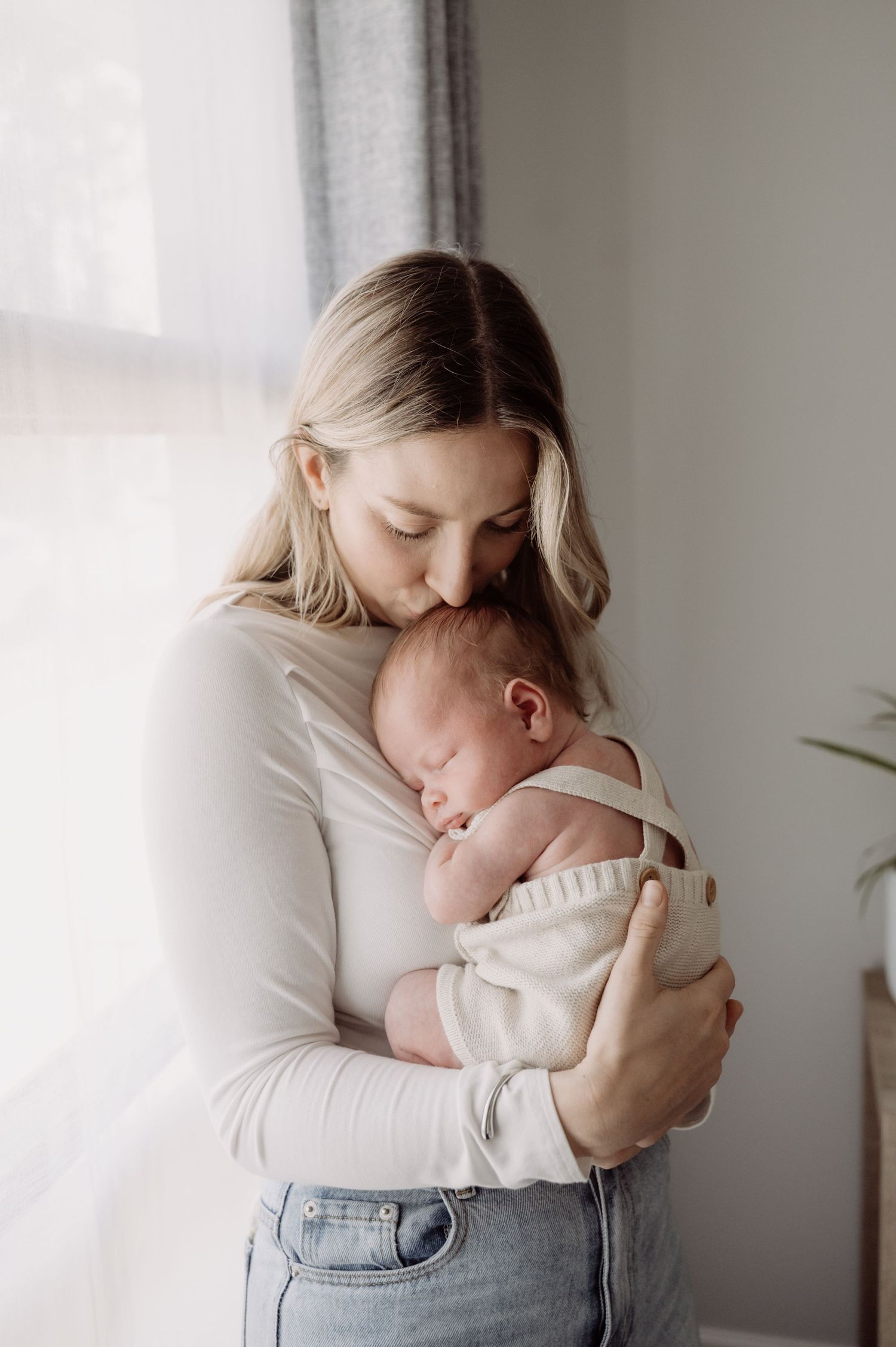 A mother tenderly kisses her sleeping newborn baby while holding them close near a bright window.