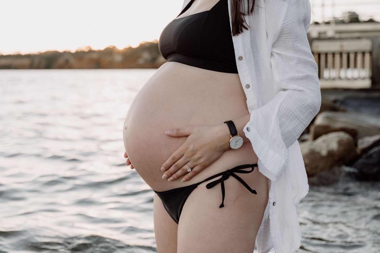 Pregnant woman in black bikini and open white shirt cradling her baby bump near a serene waterfront at sunset.