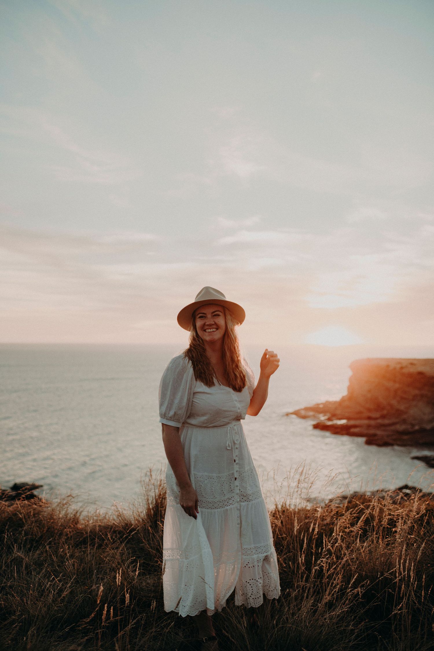 Woman in white dress and straw hat smiling at sunset on coastal cliffs.