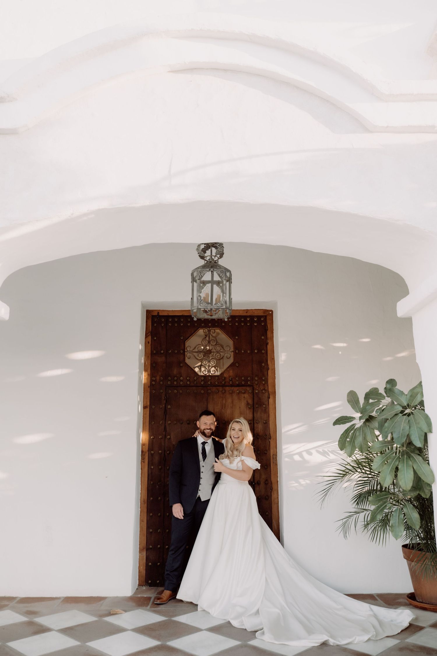 Bride and groom pose under white arched entryway with ornate wooden door and hanging lantern on checkered floor.