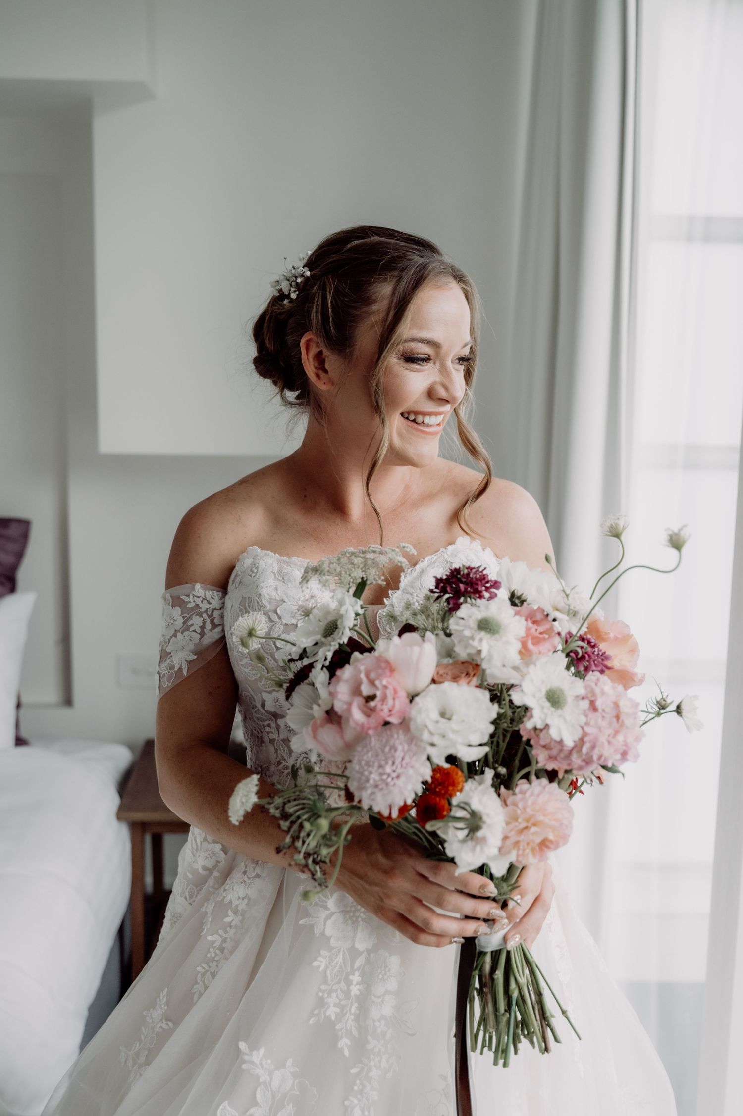 Smiling bride in off-shoulder lace gown holds lush pink and white floral bouquet near bright window.