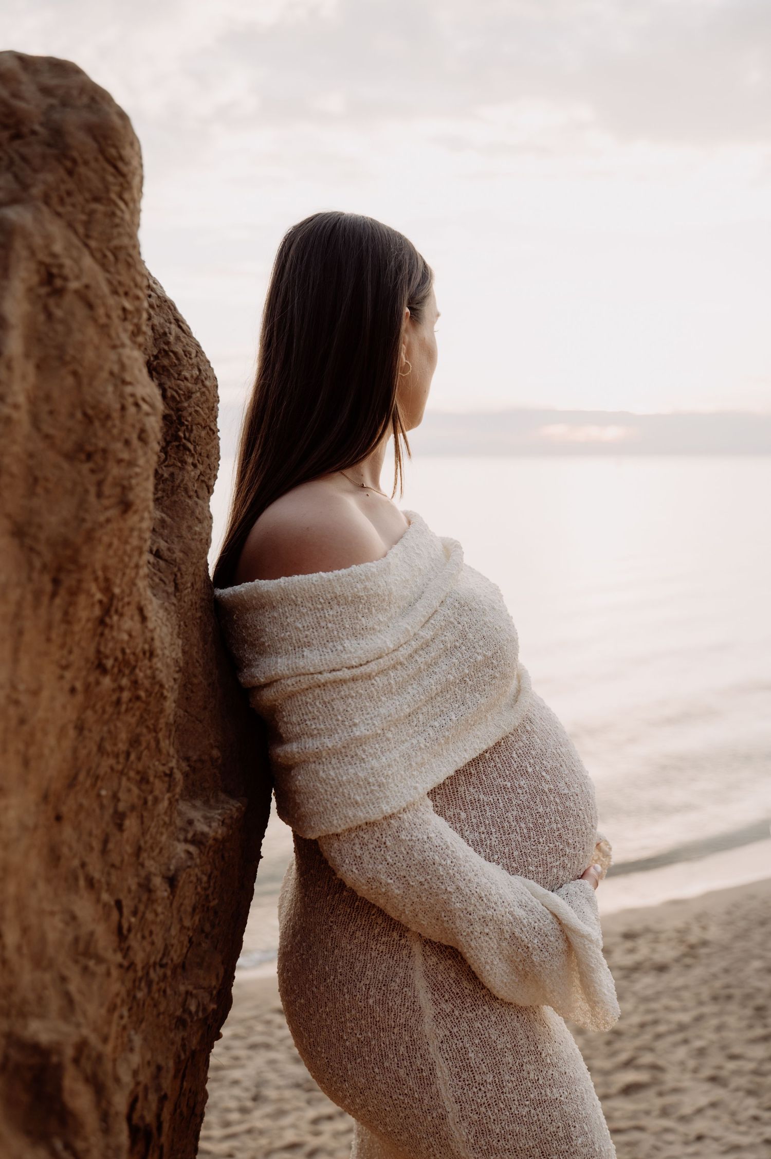 Pregnant woman in cream knit wrap dress leaning against rock on beach at sunset gazing at ocean.