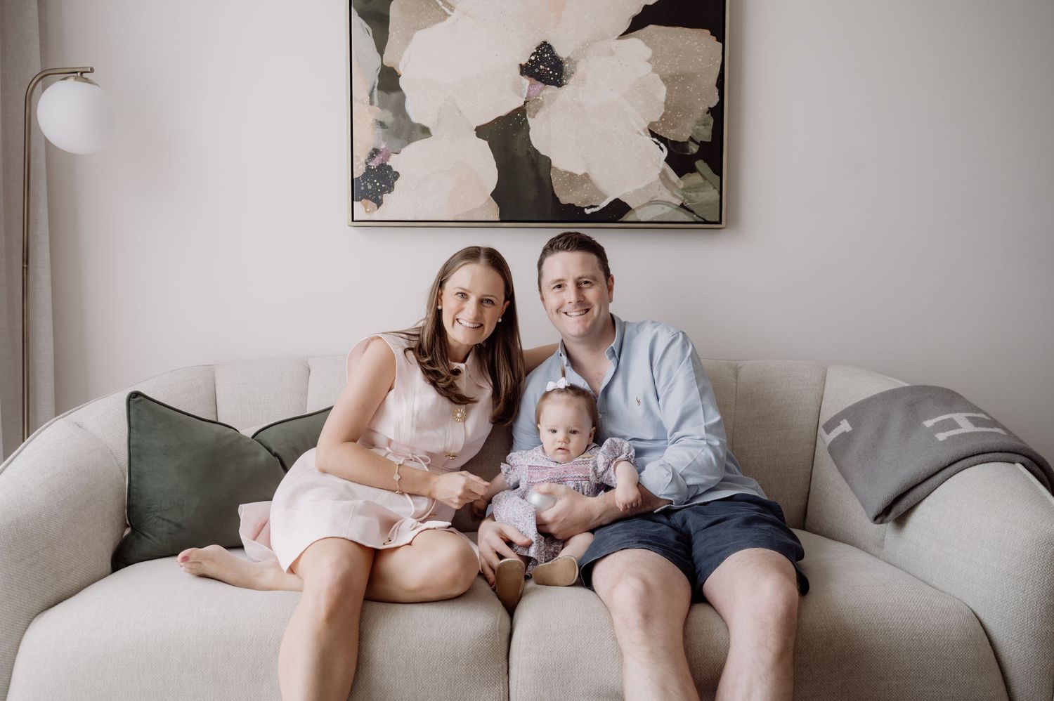Happy family of three sitting on a cream sofa, parents smiling with baby in a bright modern living room.
