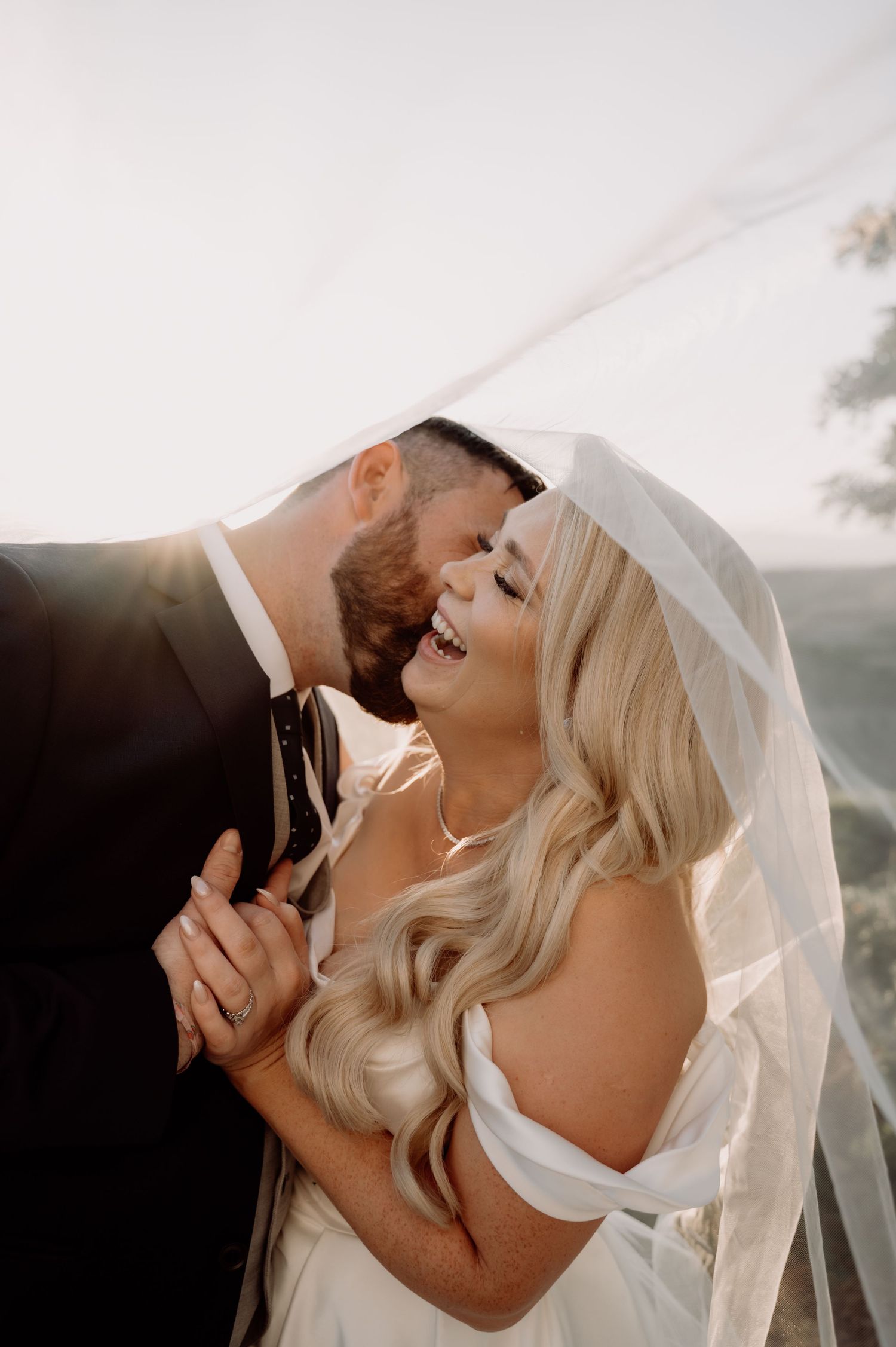 A joyful bride and groom share a romantic moment under a flowing veil, laughing together in golden wedding day sunlight.
