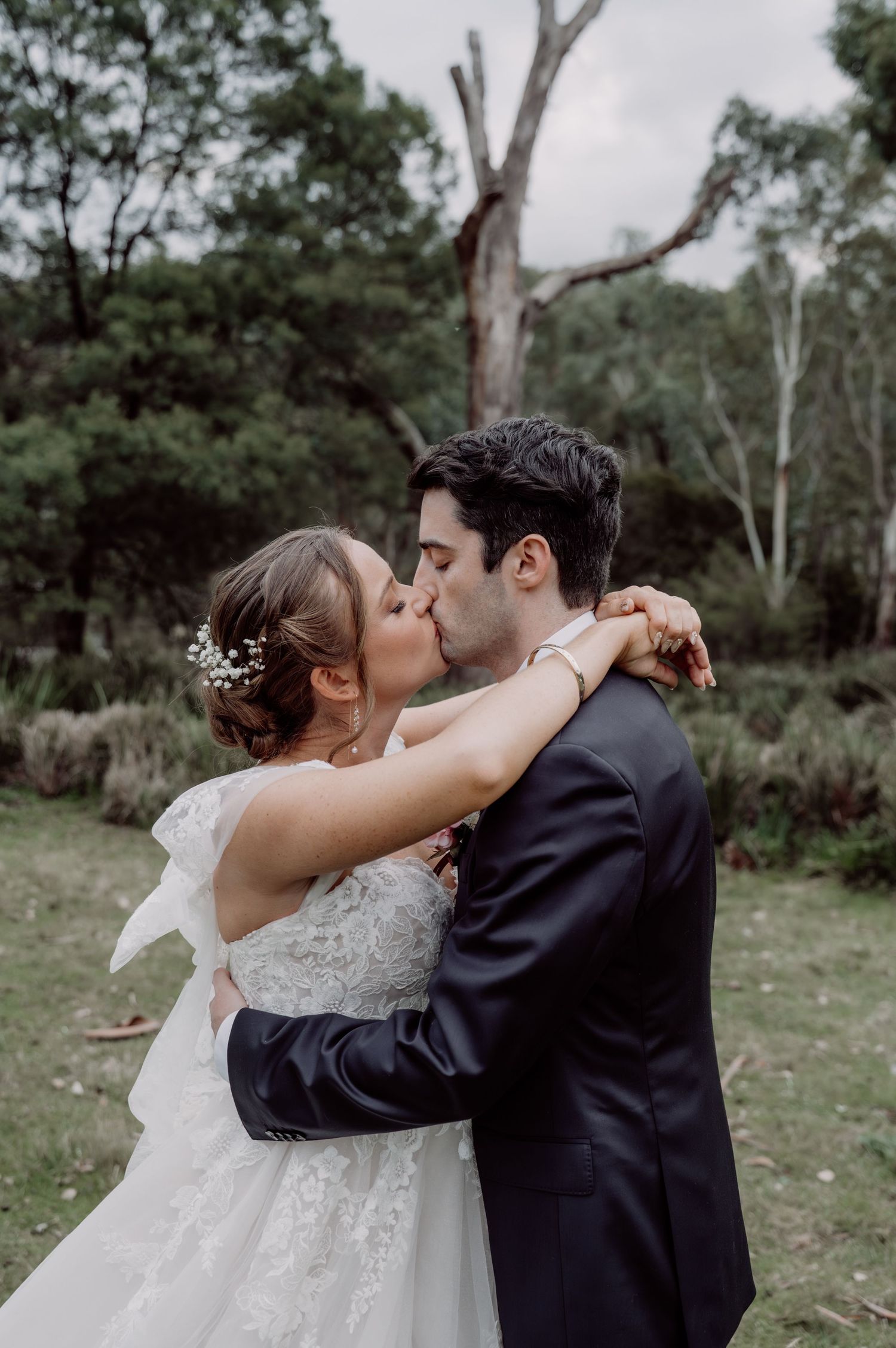 A bride and groom share a romantic kiss outdoors, surrounded by lush greenery, as she wraps her arms around his neck.