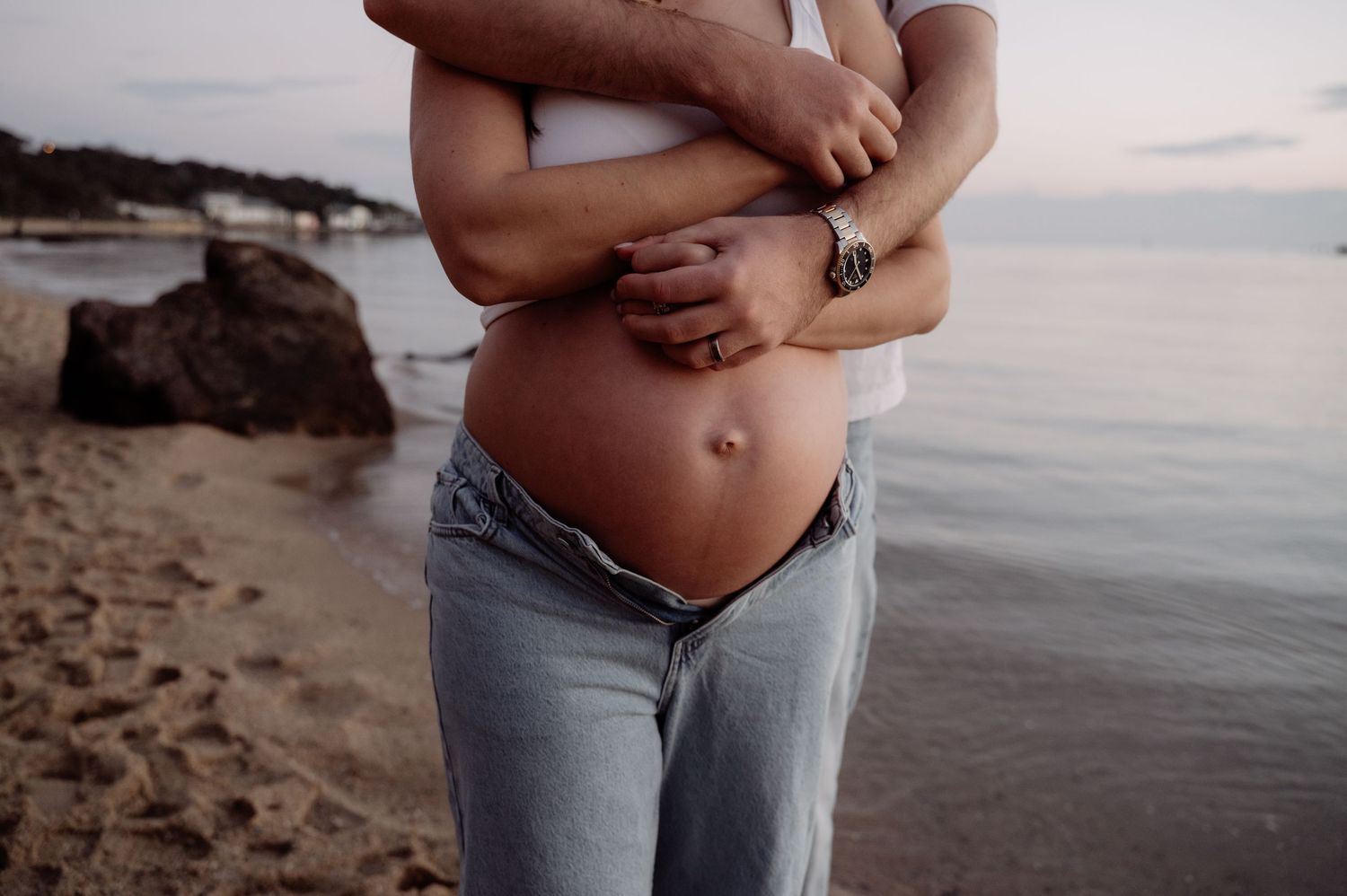 Pregnant woman in jeans embraced by partner on a sandy beach at dusk, showcasing baby bump.