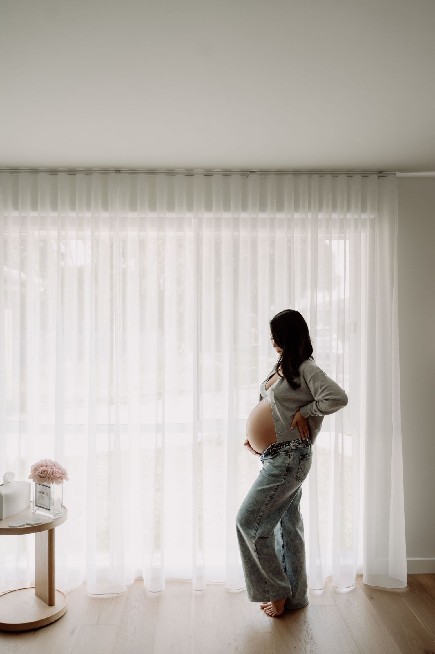 Pregnant woman standing by sheer white curtains in natural light, wearing jeans and crop top, showing baby bump.
