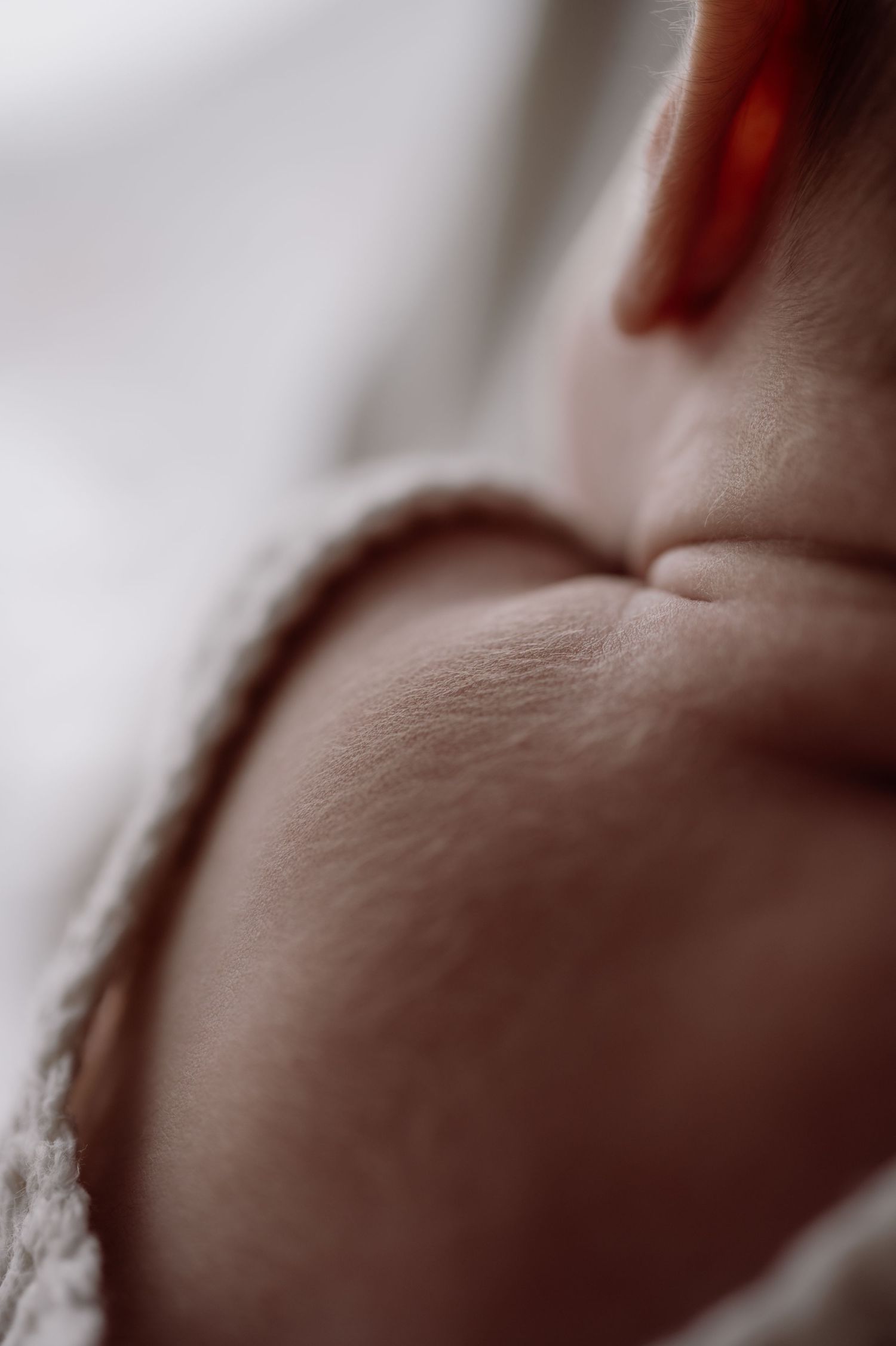 Close-up macro photo of a newborn baby's chubby chin and neck folds, with soft white fabric and an adult's red fingernail.