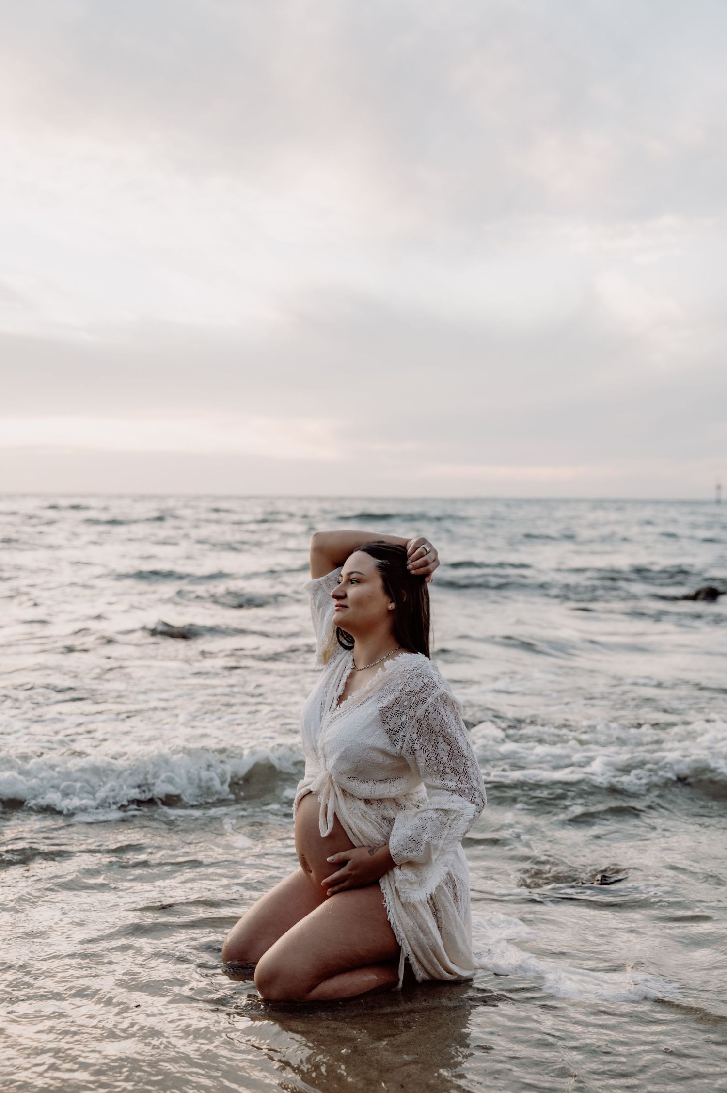 Pregnant woman in white lace robe kneeling in ocean waves at sunset, cradling baby bump.