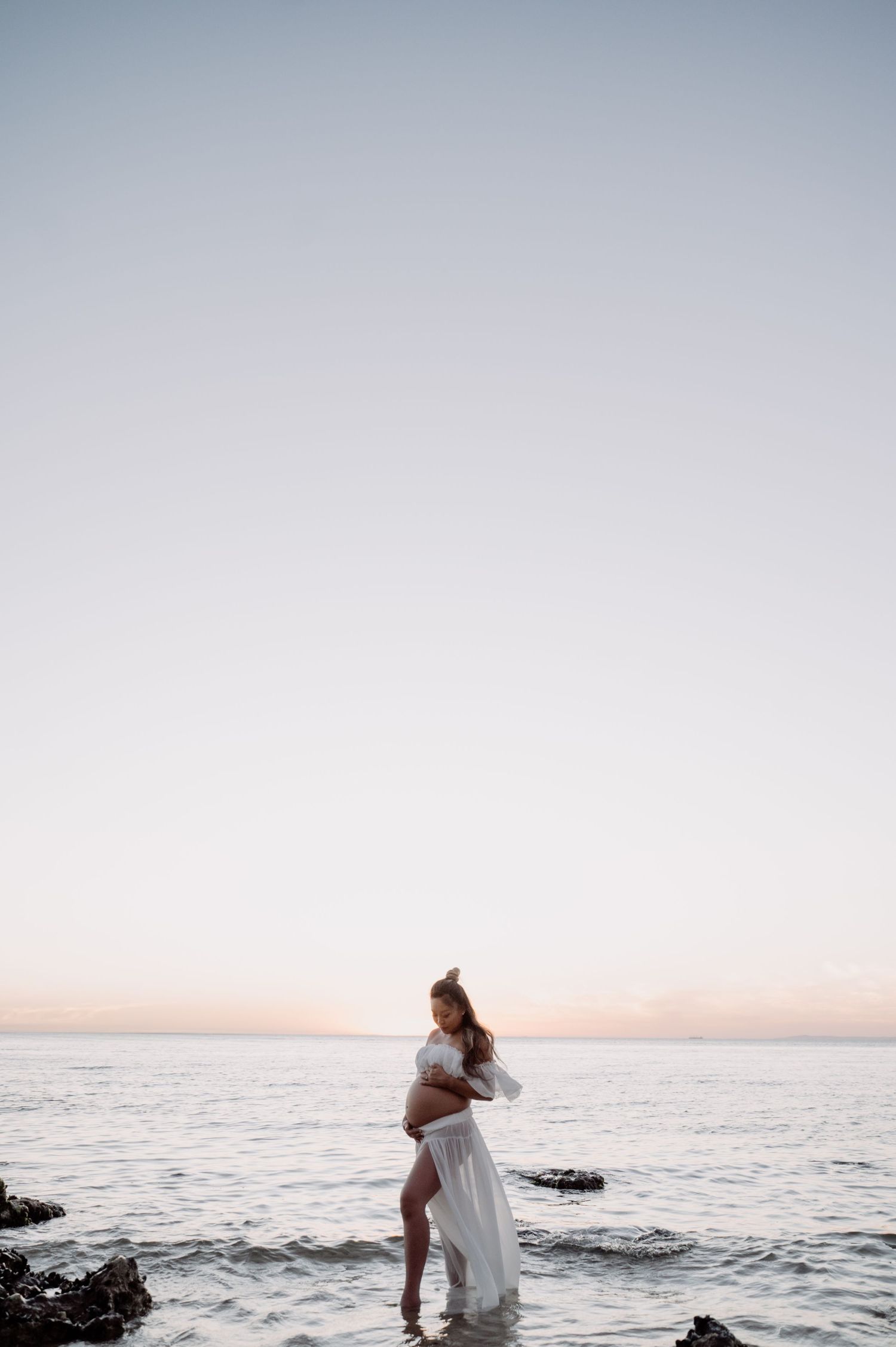 Pregnant woman in flowing white dress standing in calm ocean water at sunset.