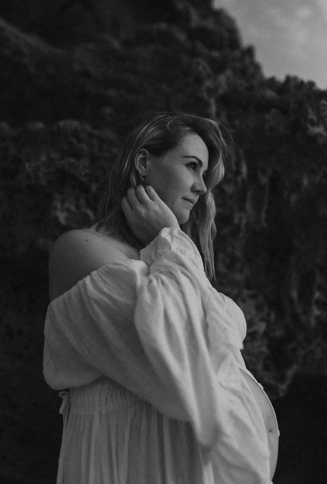 Black and white maternity portrait of pregnant woman in white off-shoulder dress outdoors.