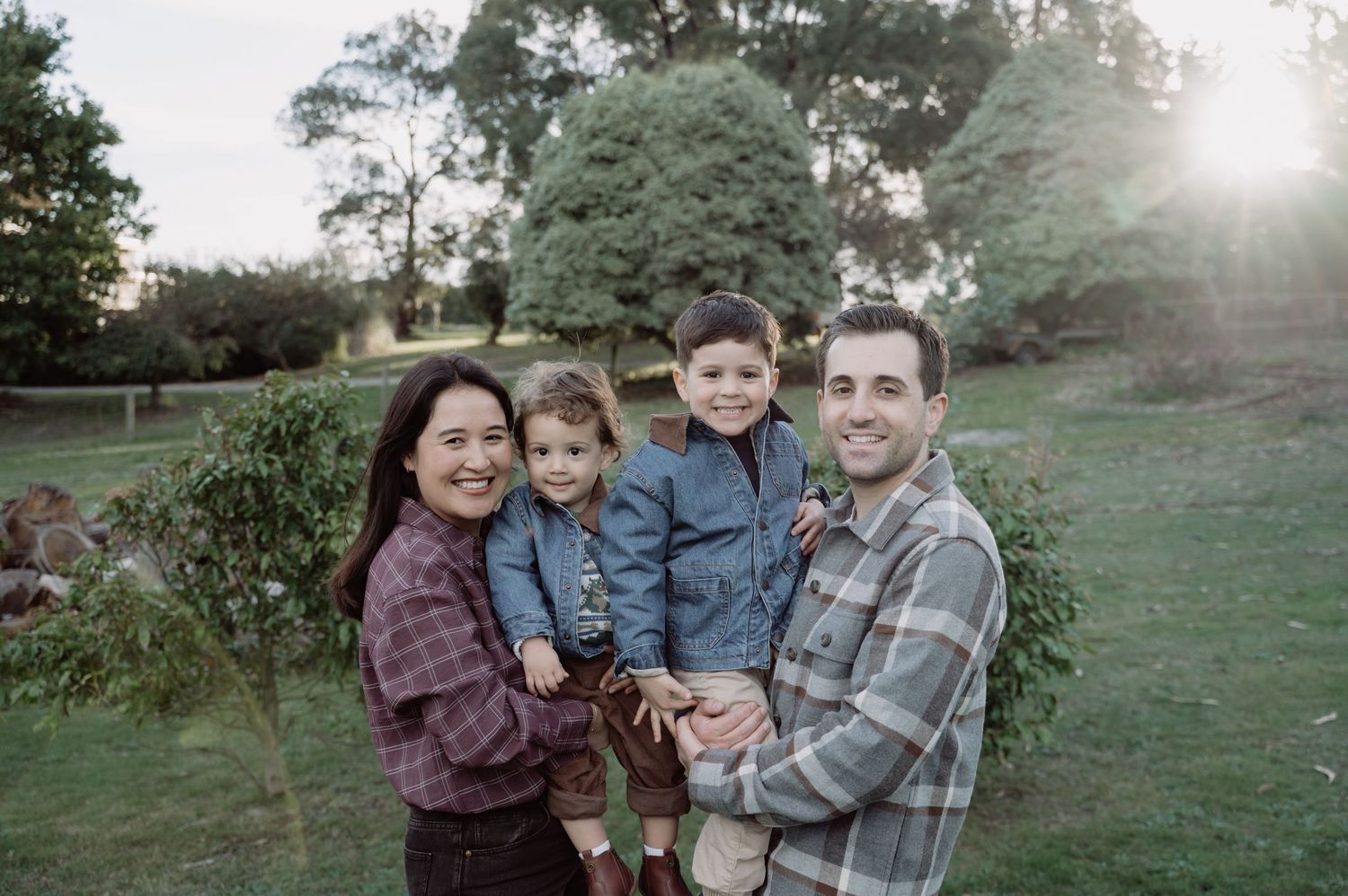 Happy family of four posing outdoors in a lush green park, parents holding two young children, all smiling warmly.