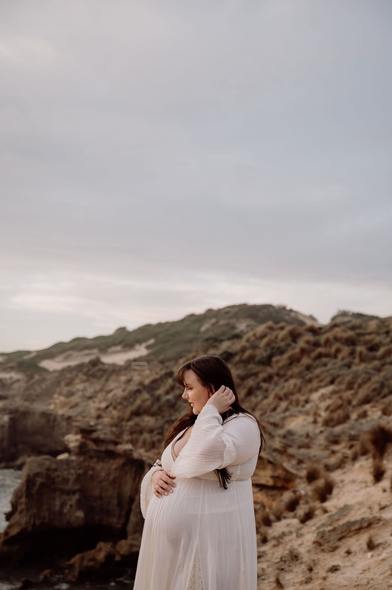 Pregnant woman in white sheer gown cradling her bump at rocky coastal cliffs under overcast sky.