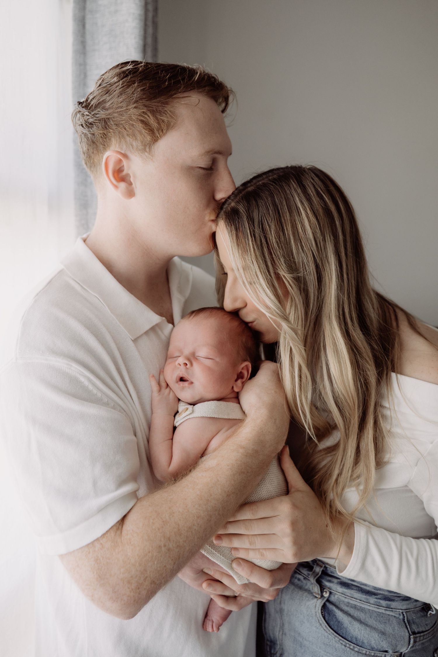 A couple tenderly kisses their sleeping newborn baby near a window in a soft, intimate family portrait.