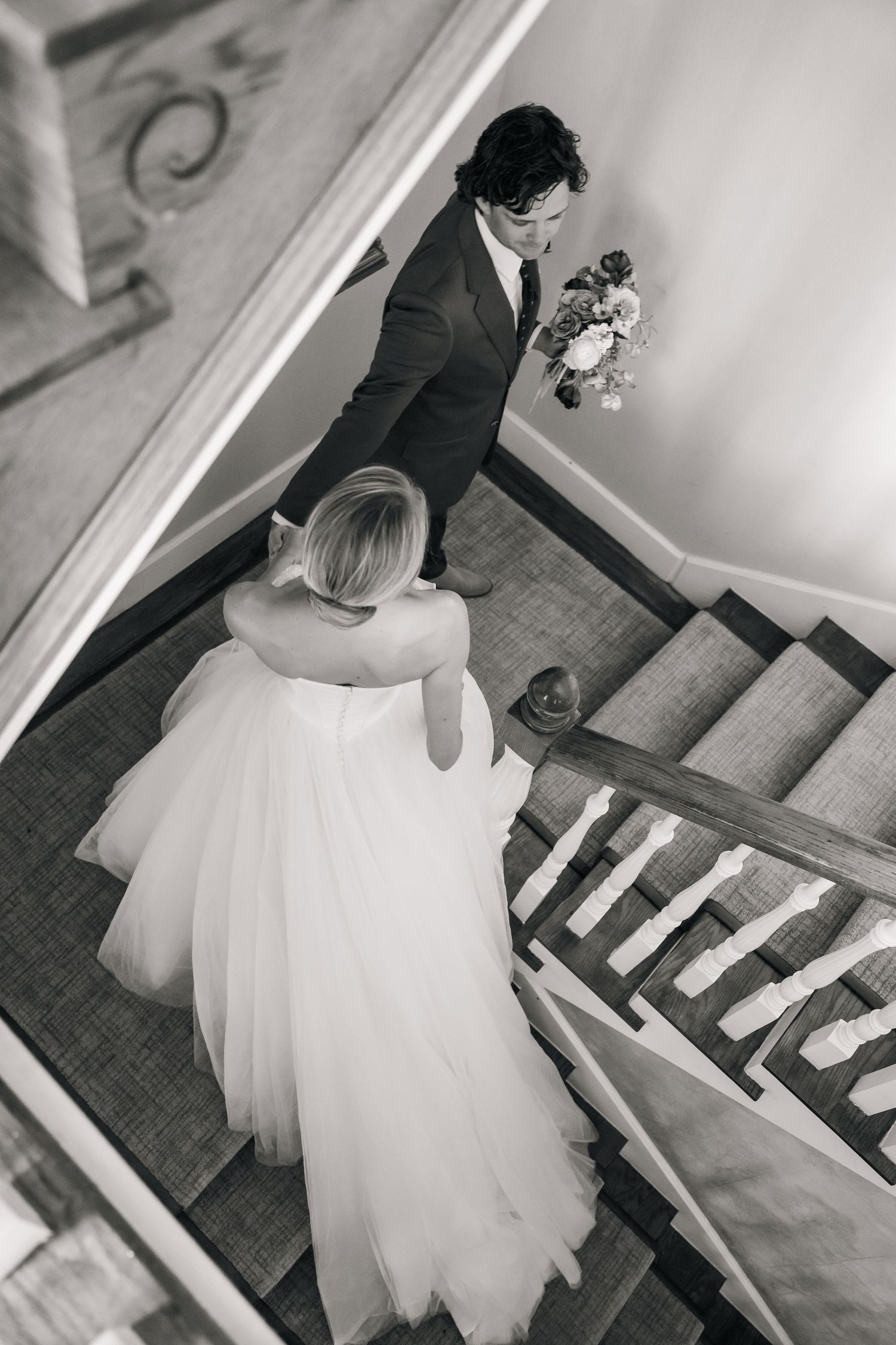 Black and white photo captures wedding couple descending vintage wooden staircase.