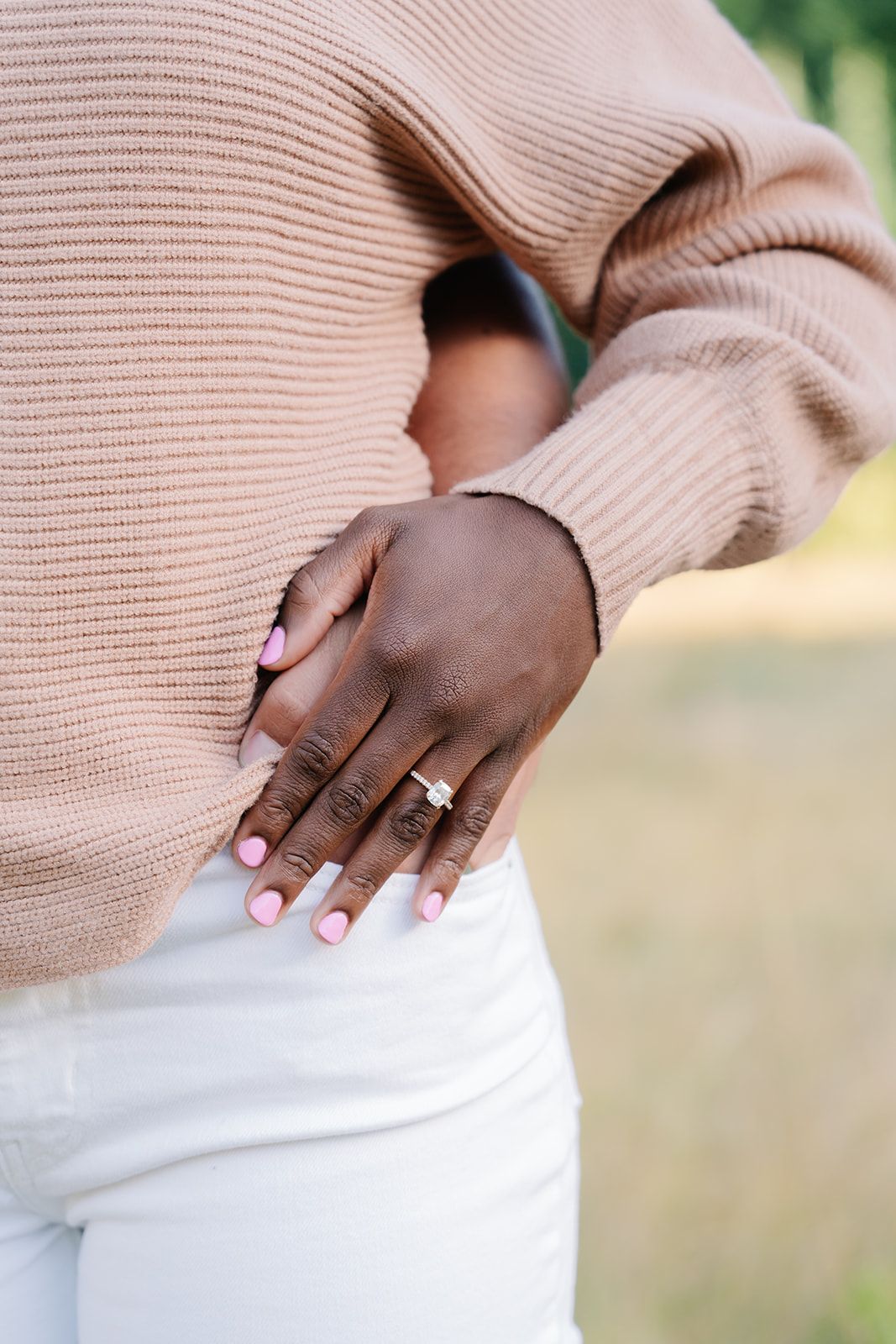 Detail shot of hands with engagement ring against soft pink sweater and white clothing.