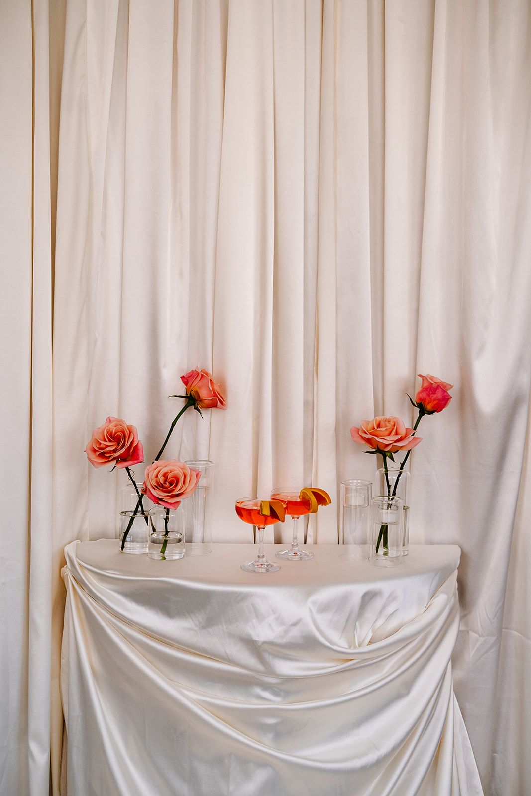 Coral roses in glass vases and small cocktails arranged on a white draped table against a light backdrop.