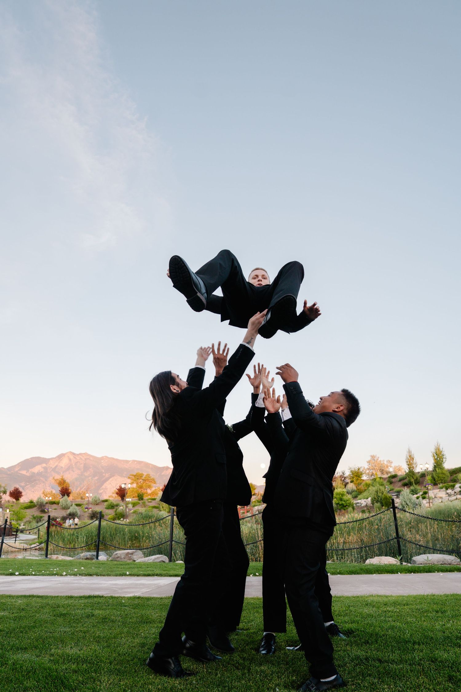 Group of friends in black suits tossing a person into the air outdoors with mountain backdrop.