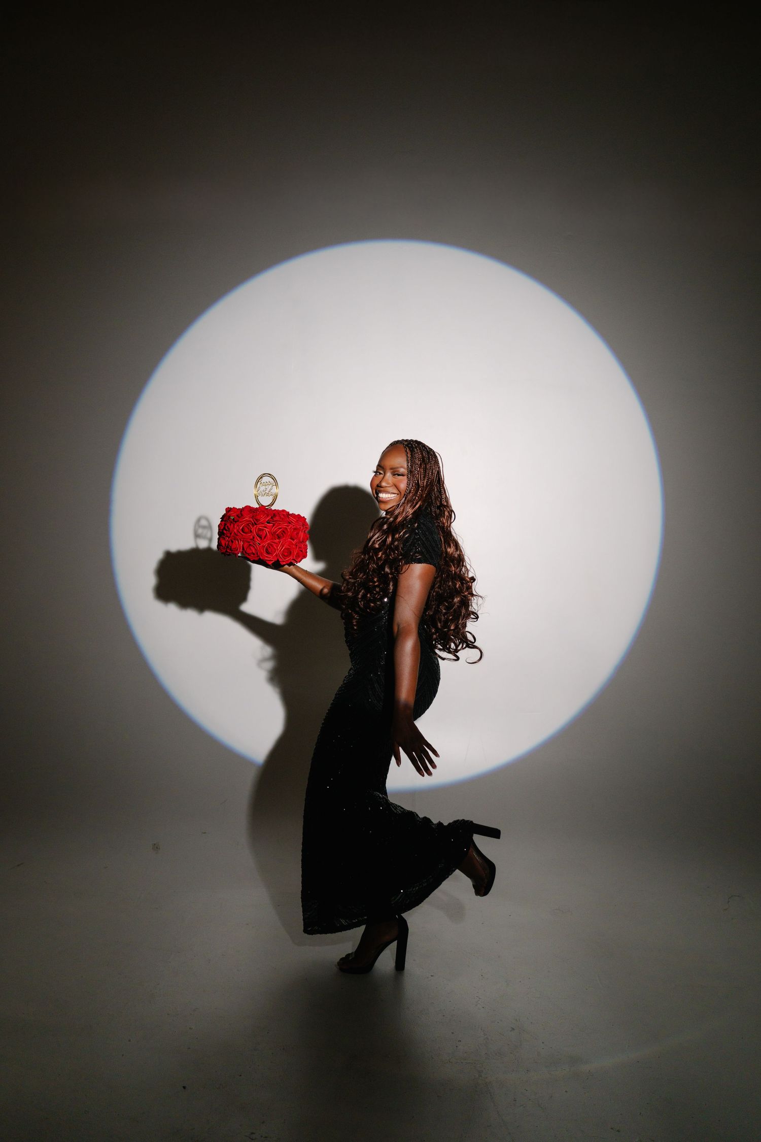 A joyful woman in a black dress holds a red rose cake, posing in a spotlight against a gray background.