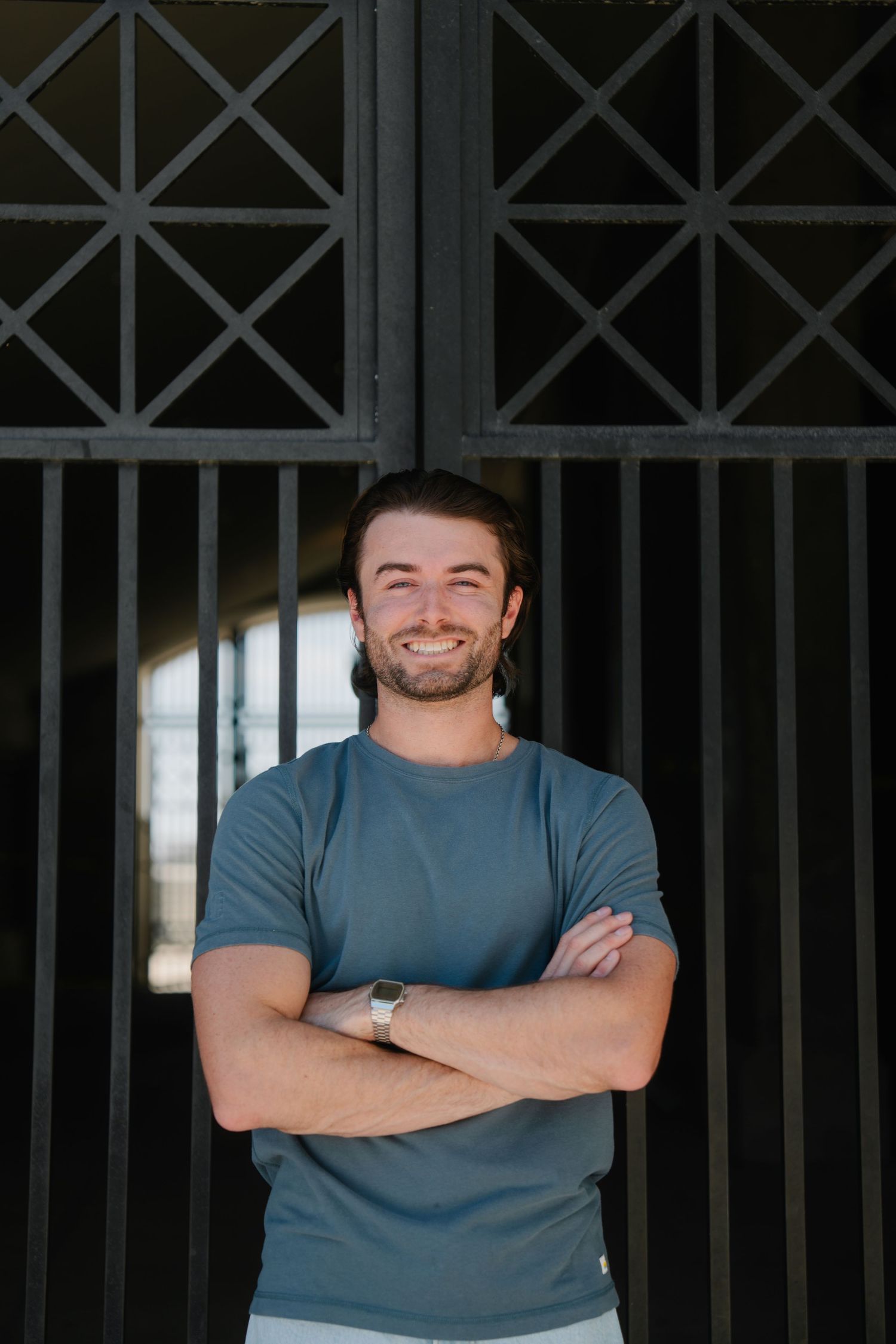 Smiling young man with beard, arms crossed, wearing blue t-shirt and watch, standing in front of dark metal gate.