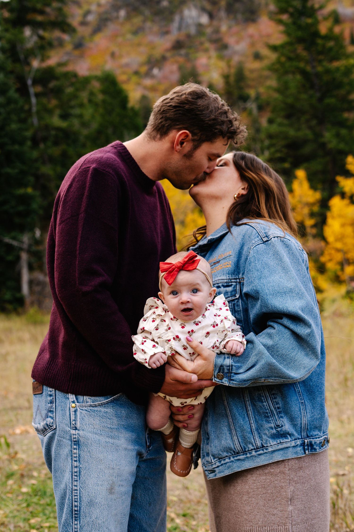 A couple kisses while holding their baby girl with a red bow outdoors among fall foliage.
