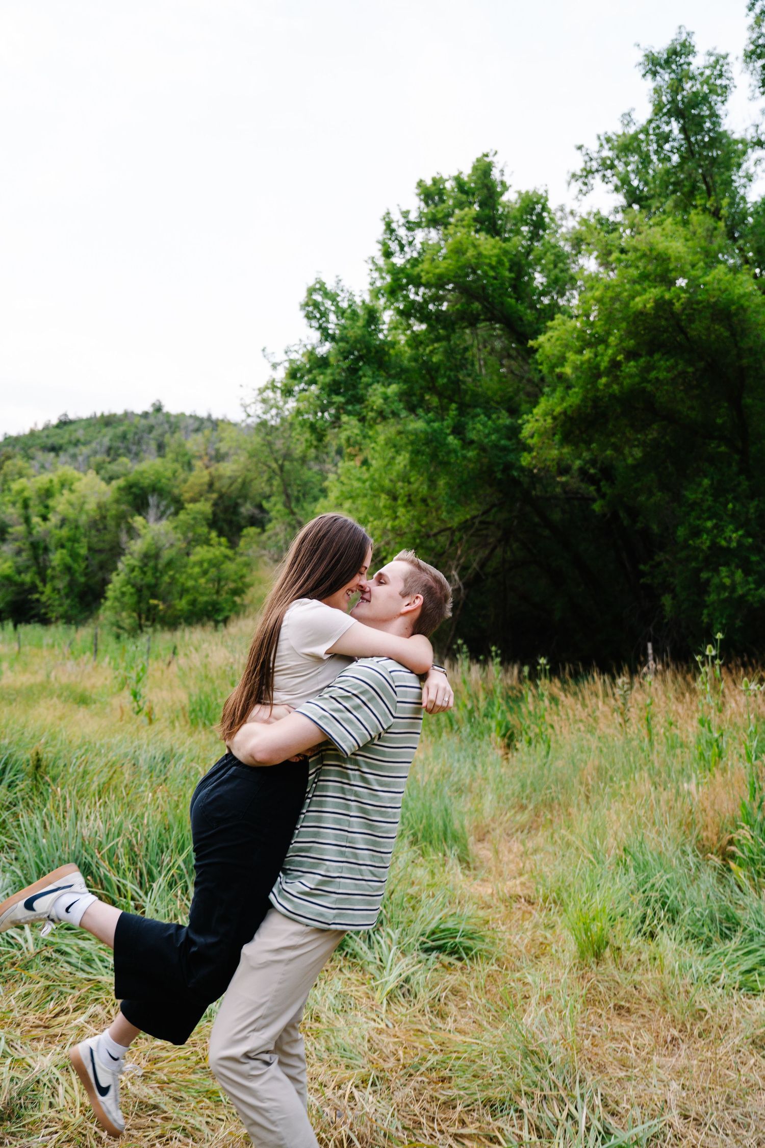 Two people share an intimate moment in a grassy field surrounded by trees.