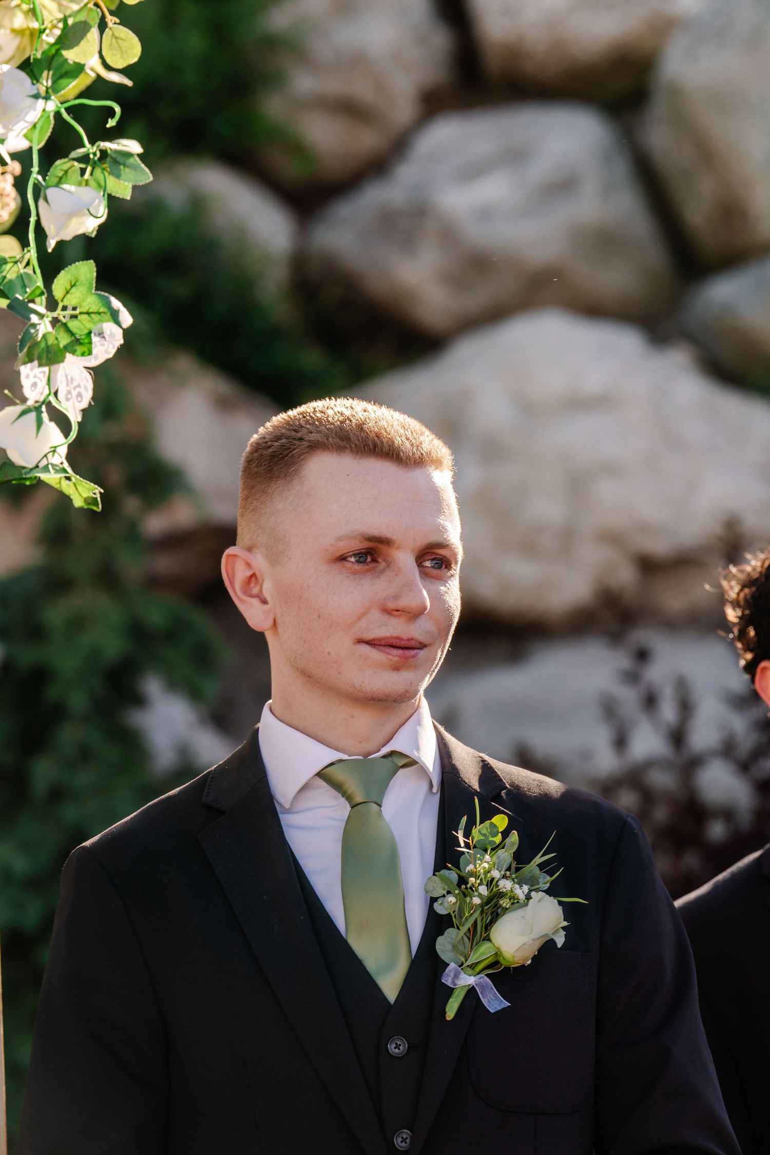 Groom wearing black suit with green tie stands near stone wall.
