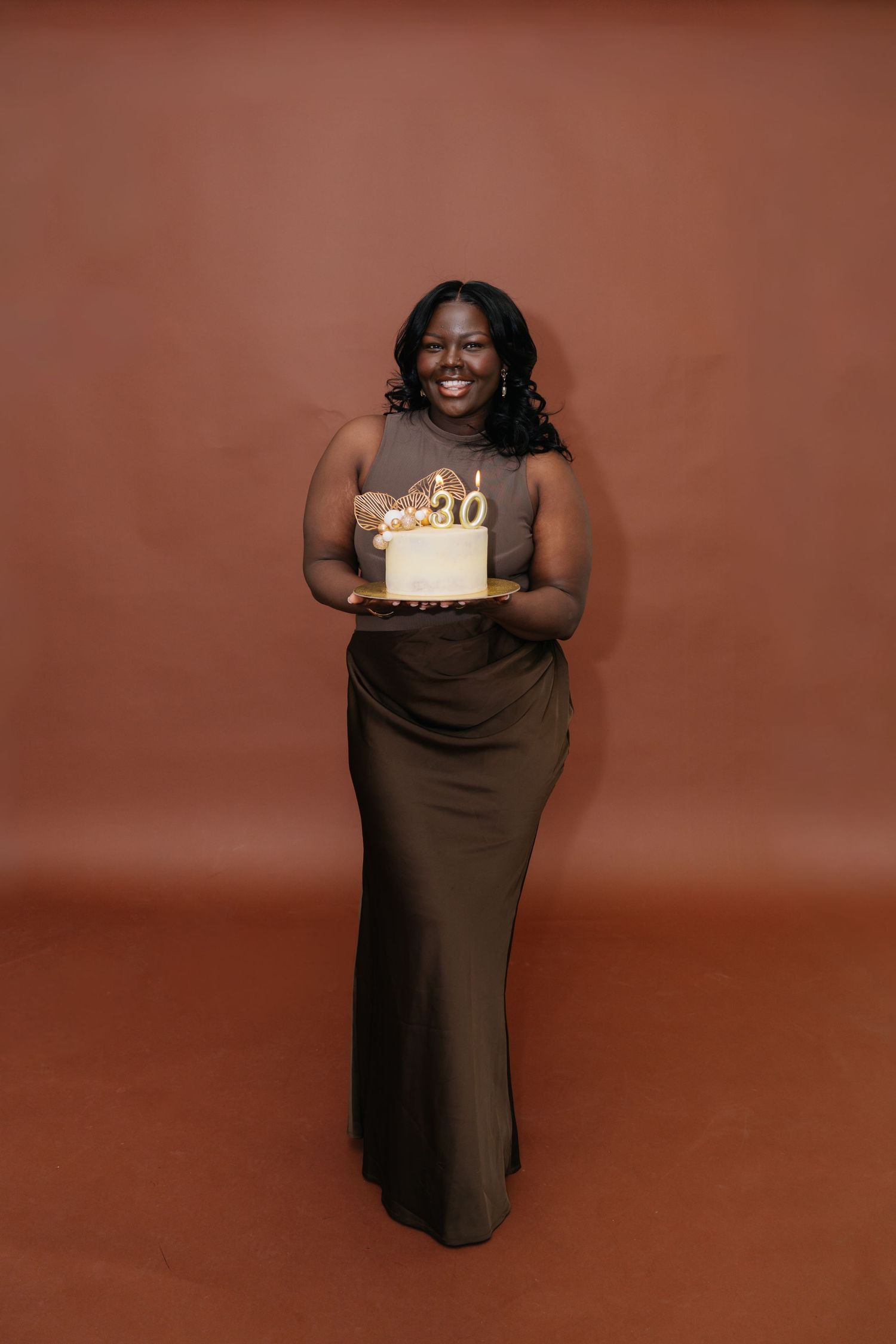 A smiling woman in an olive gown holds a white and gold 30th birthday cake against a brown backdrop.