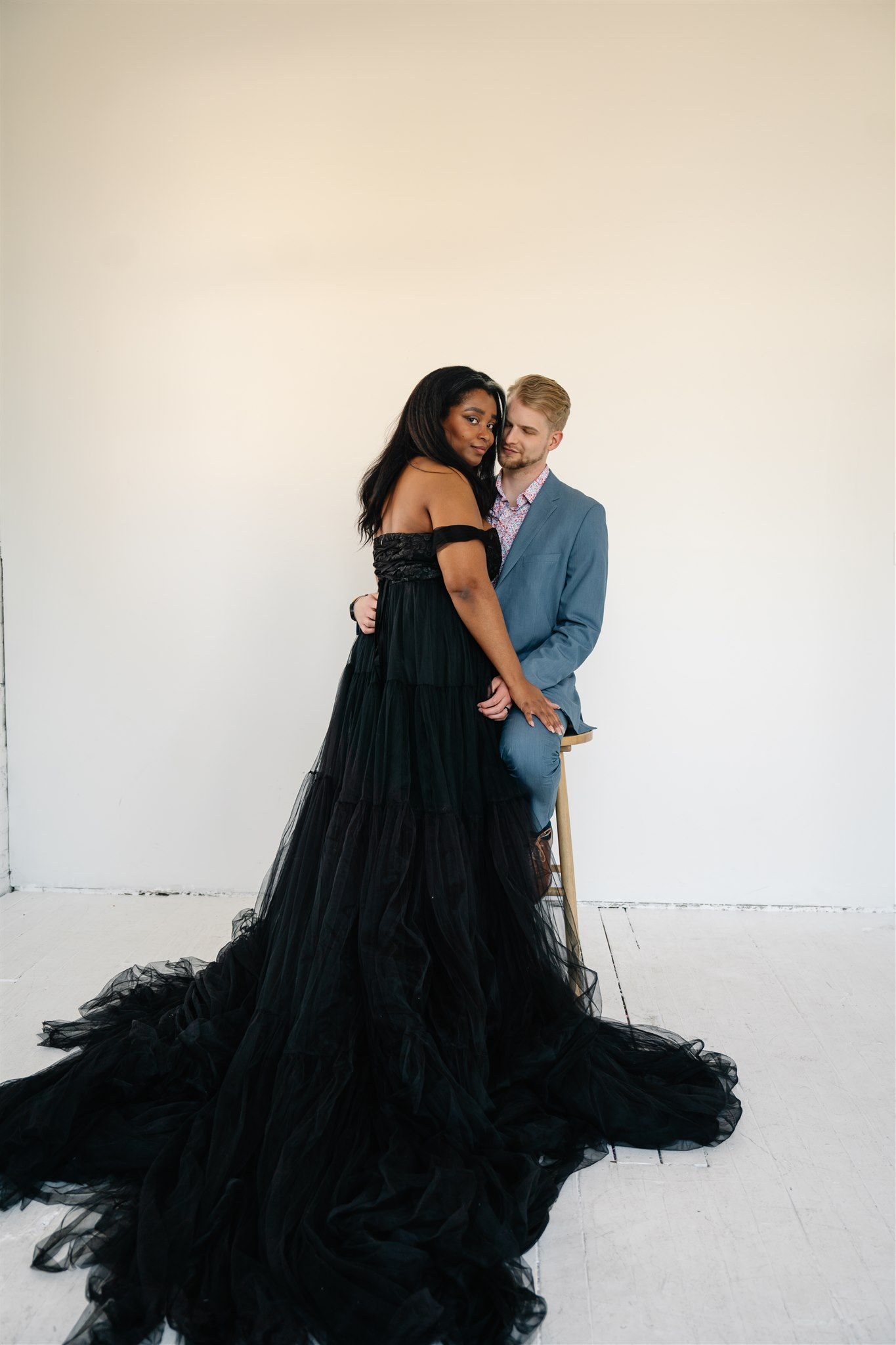 Romantic couple embracing in studio setting with dramatic black tulle gown against white wall.