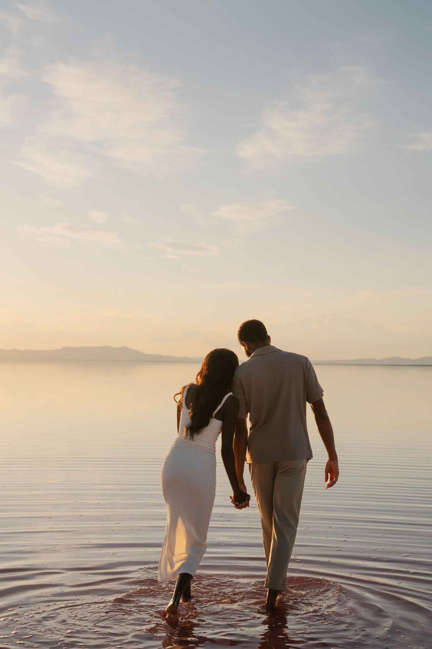 A couple wades into calm waters at sunset creating ripples in their white attire.
