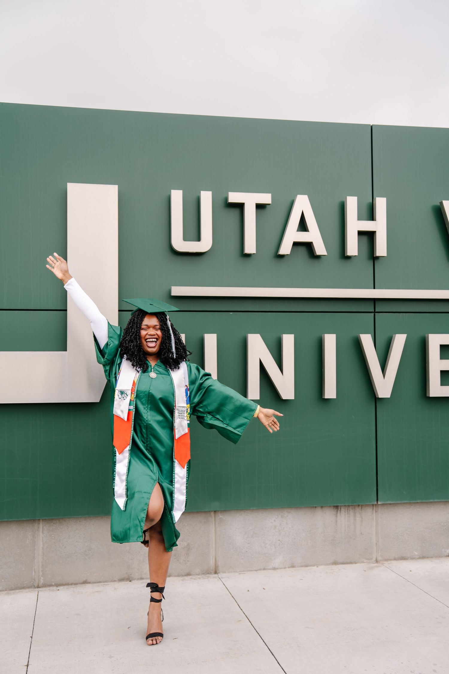 Graduate in green gown poses playfully against Utah Valley University wall signage.