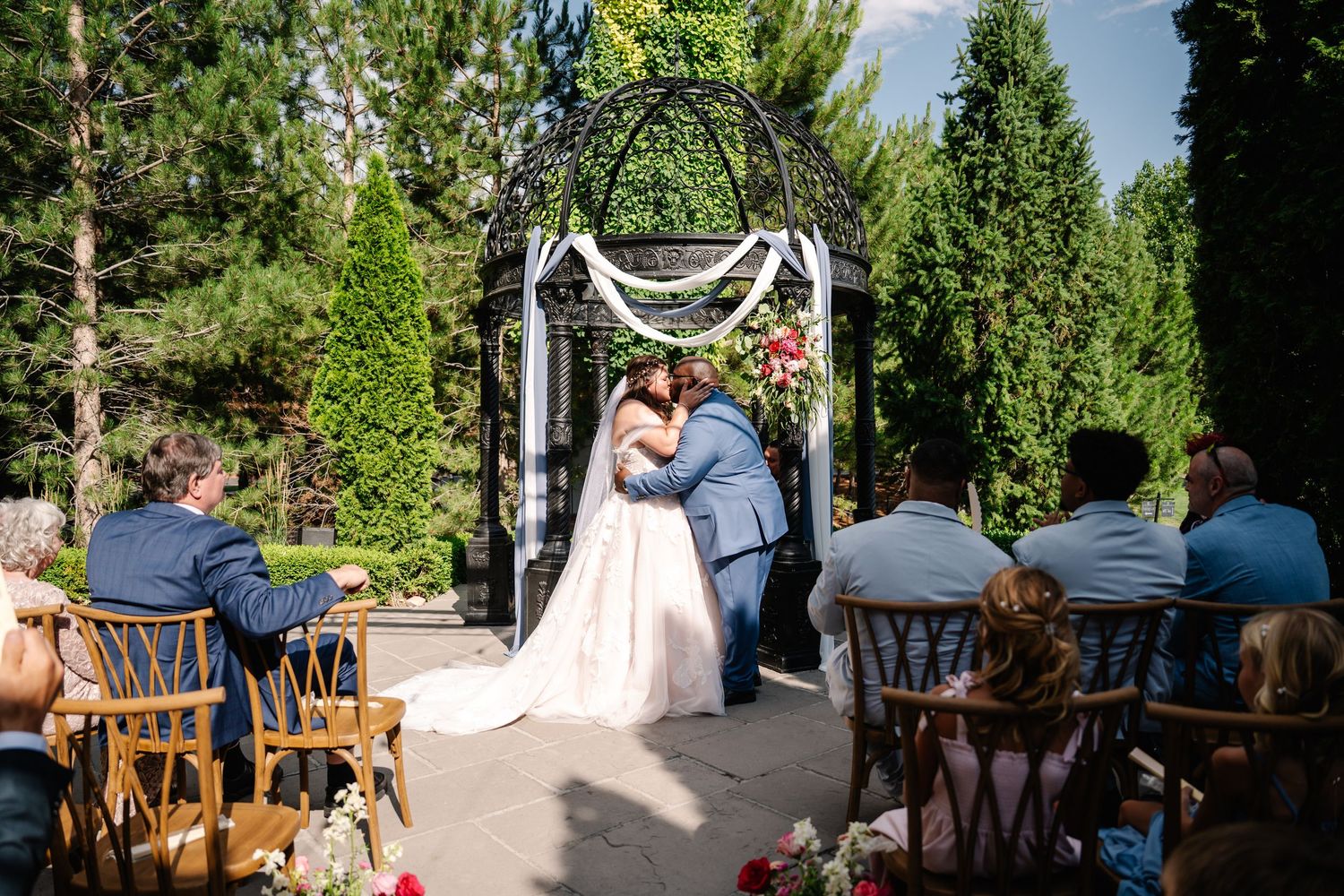 Outdoor wedding ceremony under a decorated arch with guests seated in wooden chairs surrounded by greenery.