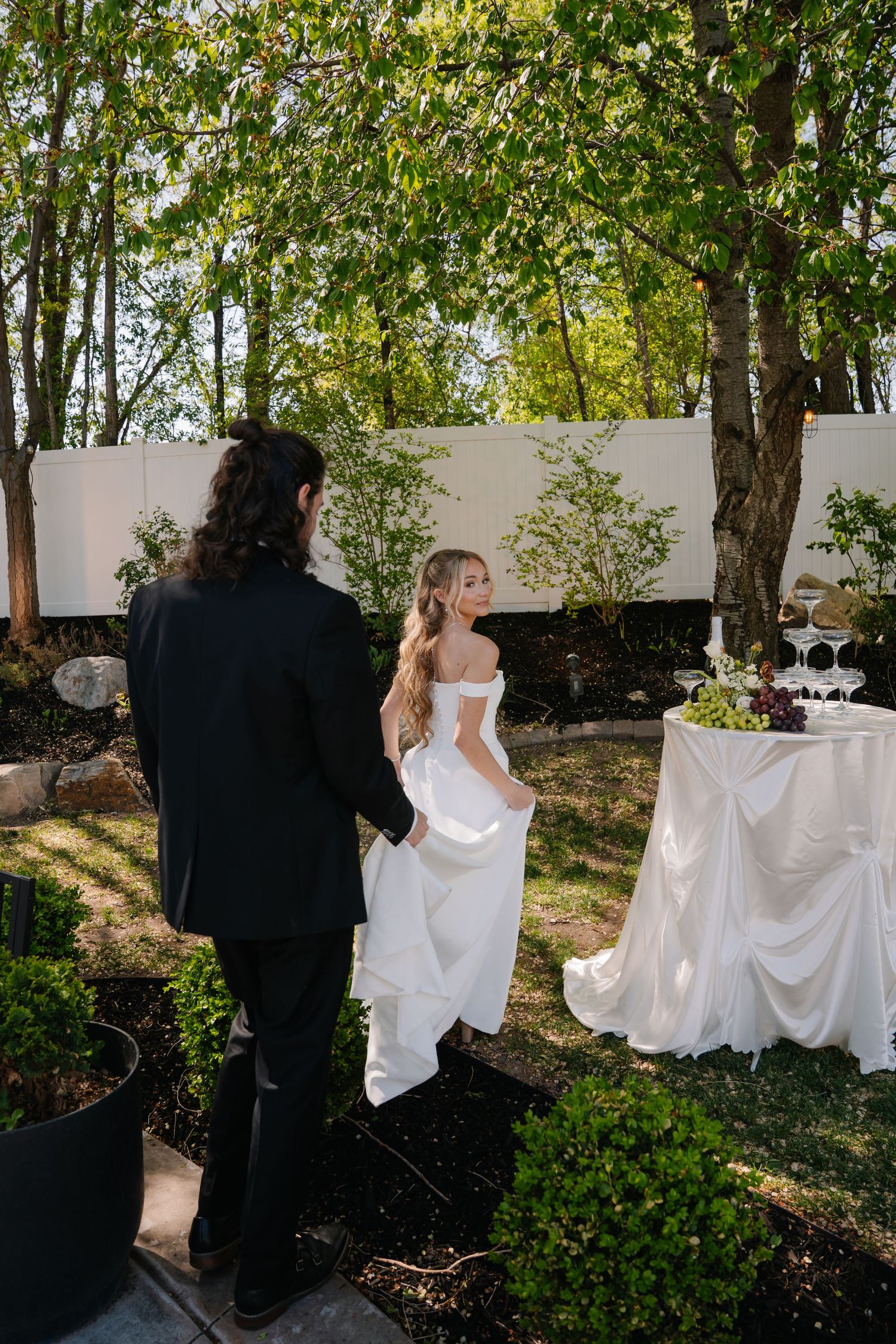 Bride in white off-shoulder gown holds hands with groom in black suit at outdoor garden wedding ceremony.