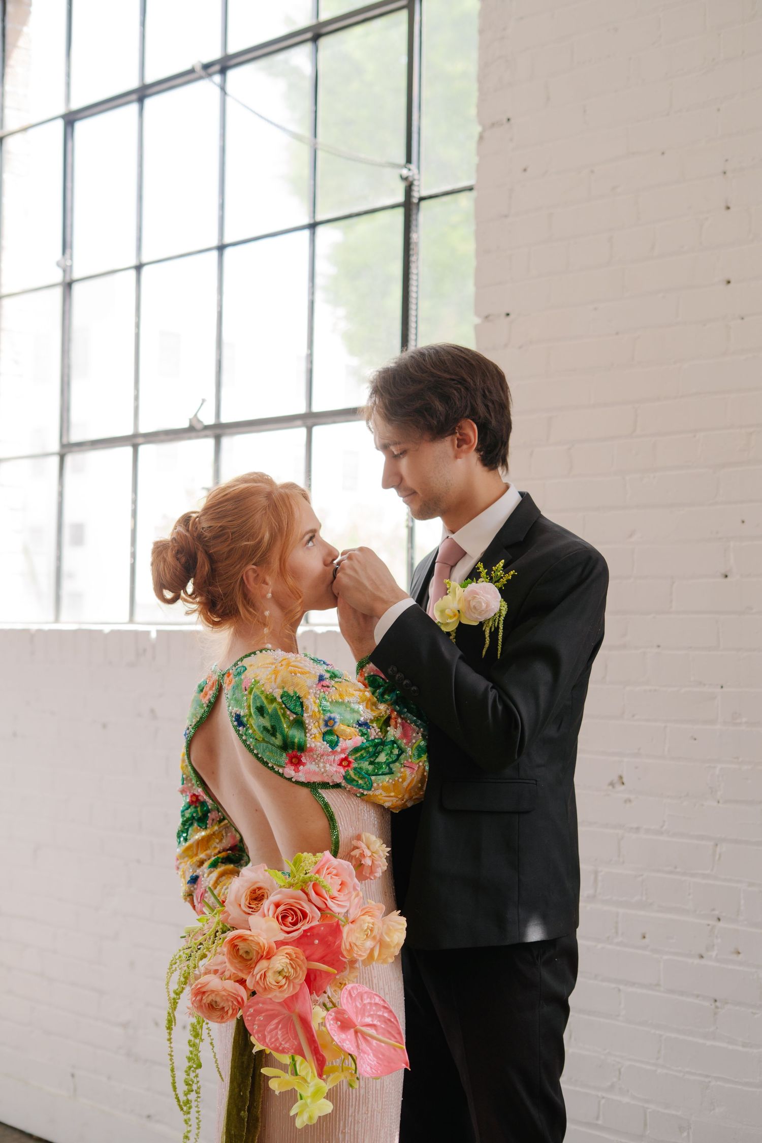 Couple in formal attire sharing intimate moments in bright industrial space with white brick walls.