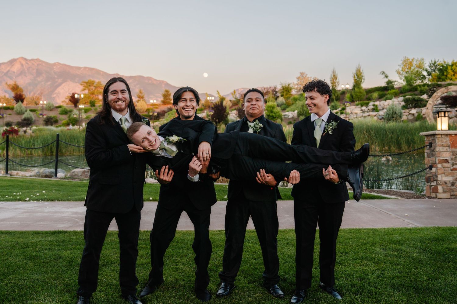 Groomsmen in black suits striking fun poses outdoors with mountains in the distance.
