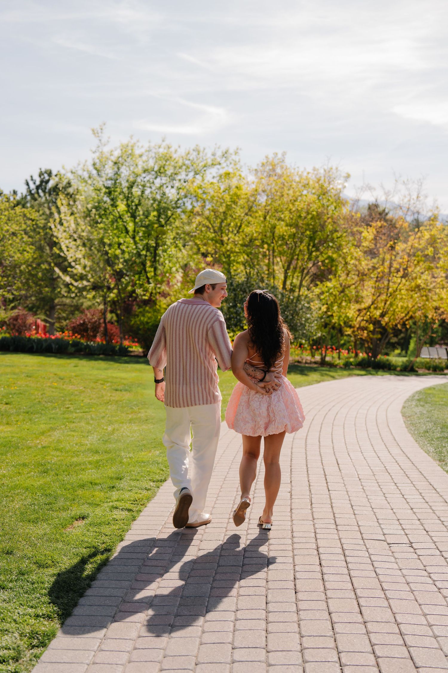 Couple walking hand in hand on a brick path in a sunny park with lush green trees and grass.