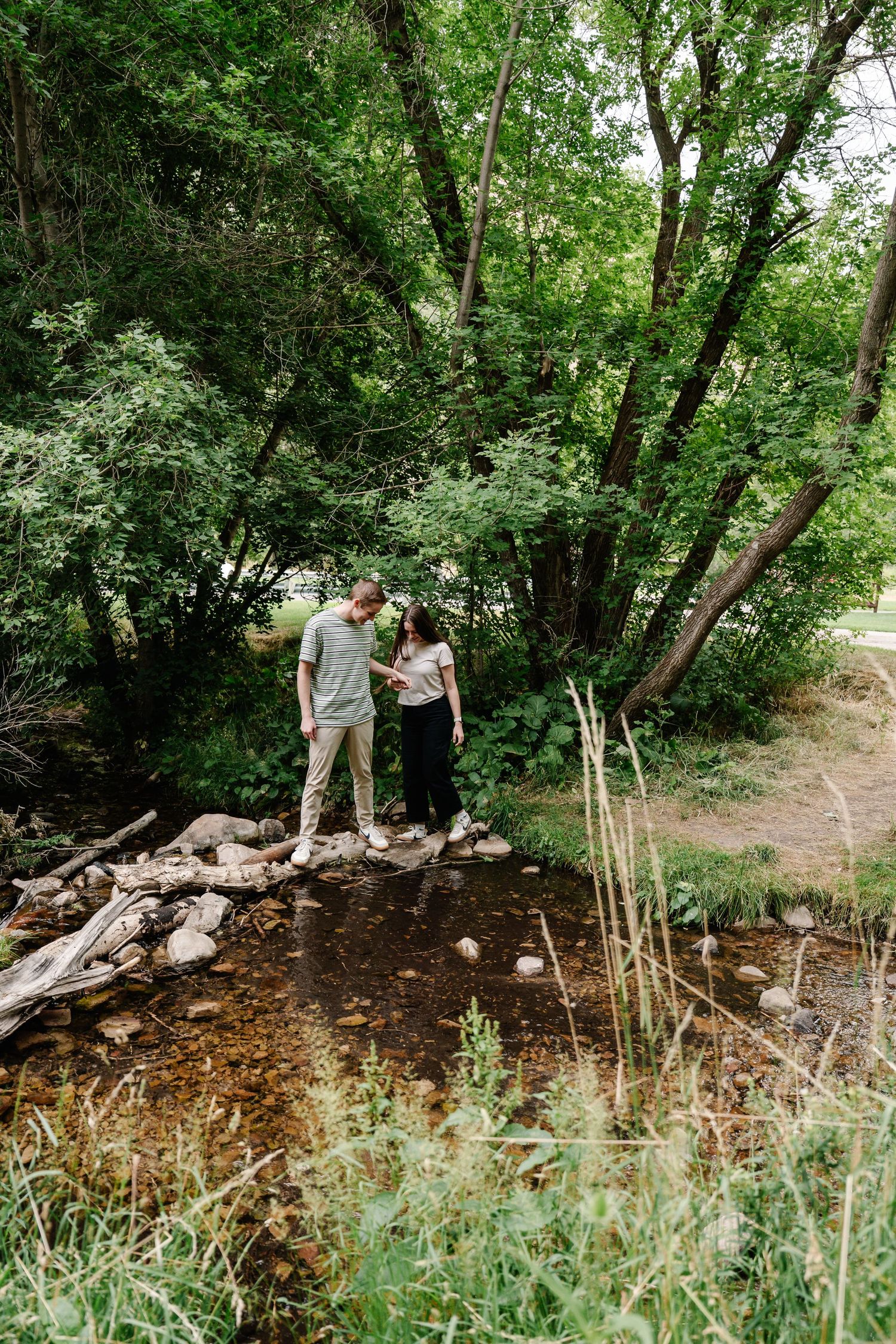 Two people explore a wooded area with rocks and tall grass.