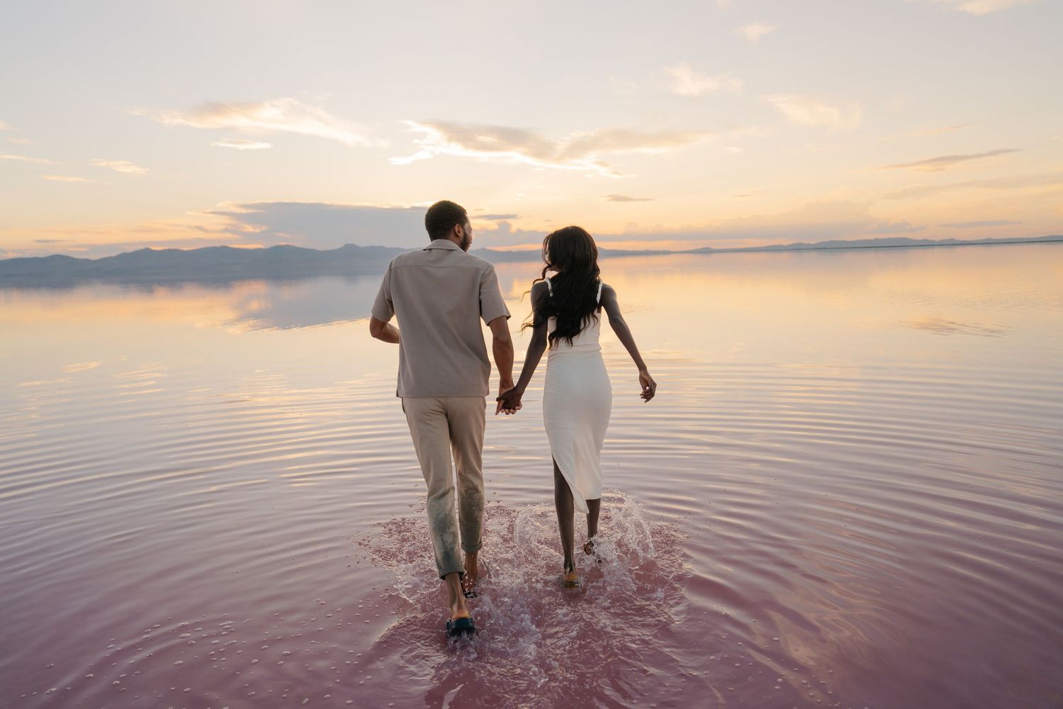 Two people splash and play in pink-colored shallow waters during sunset.
