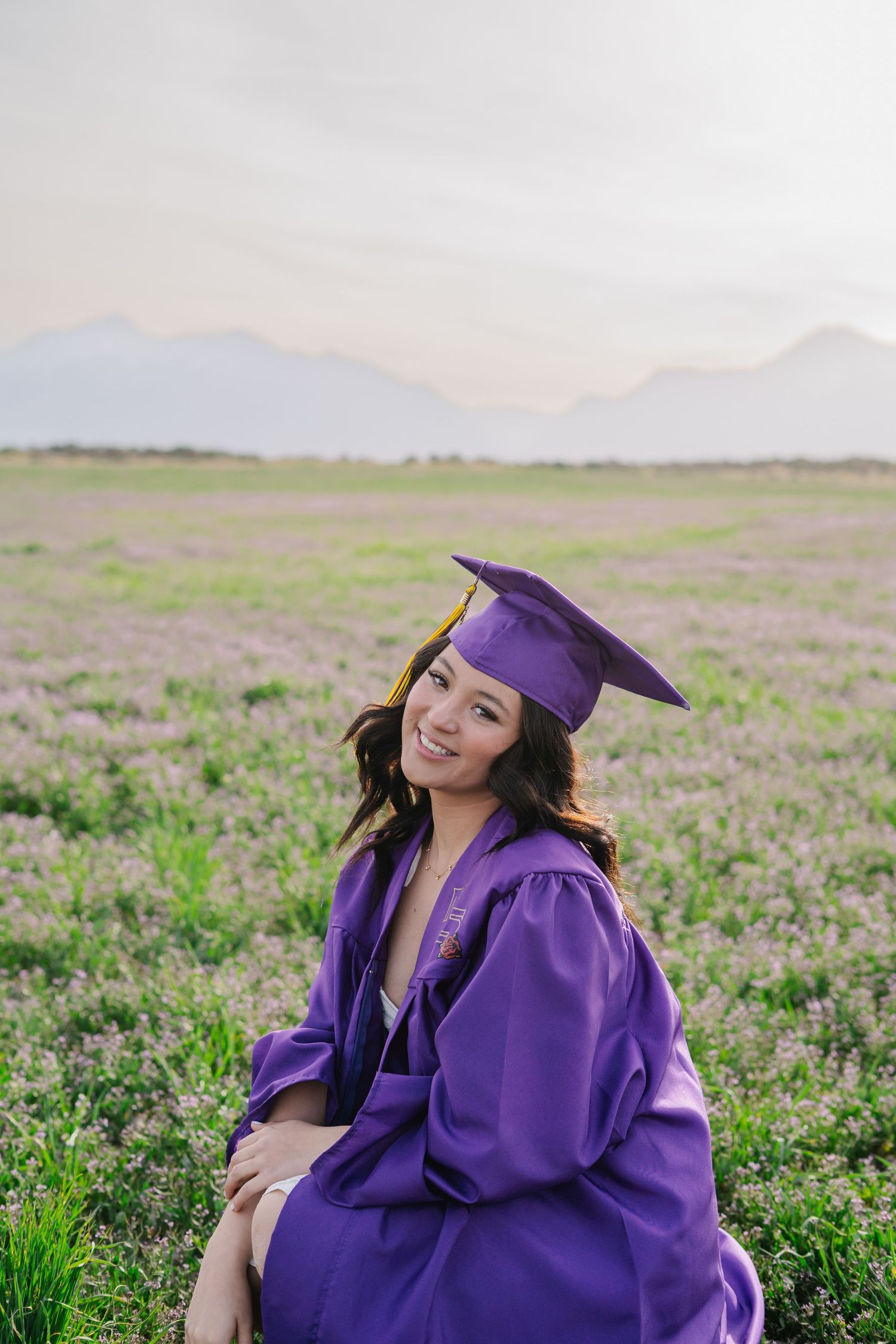 Purple graduation gown worn in a flowering meadow with mountains in background.