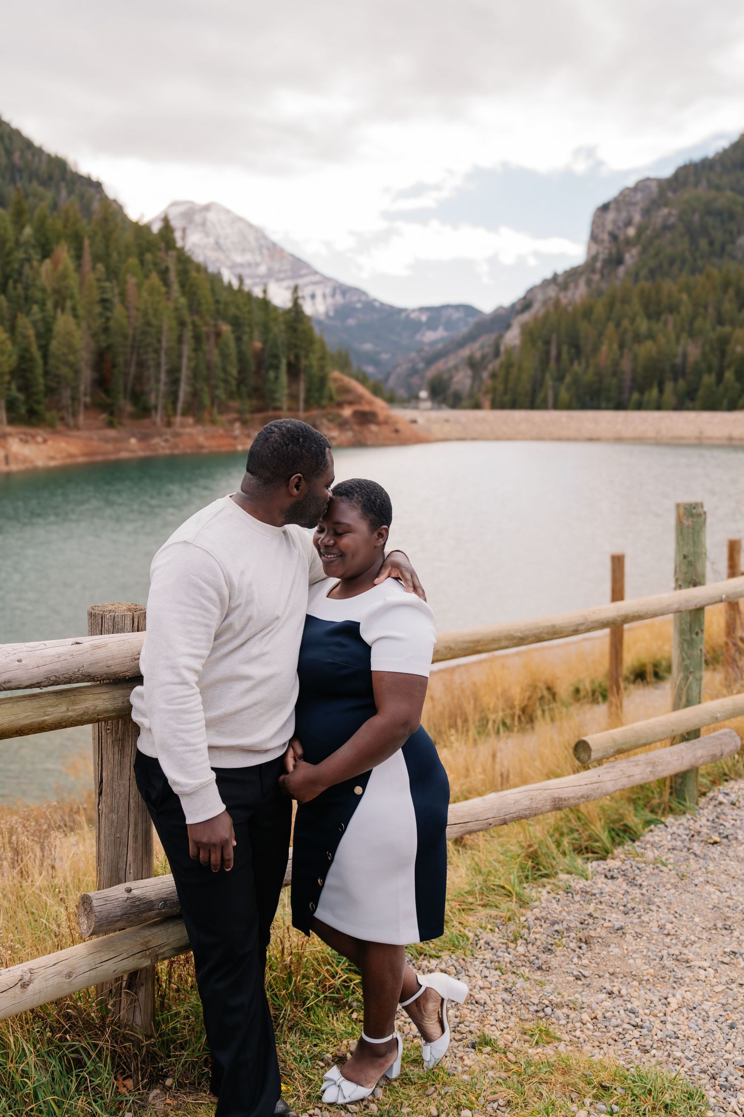 Couple embracing by a wooden fence with a turquoise mountain lake and pine forest backdrop on a cloudy day.