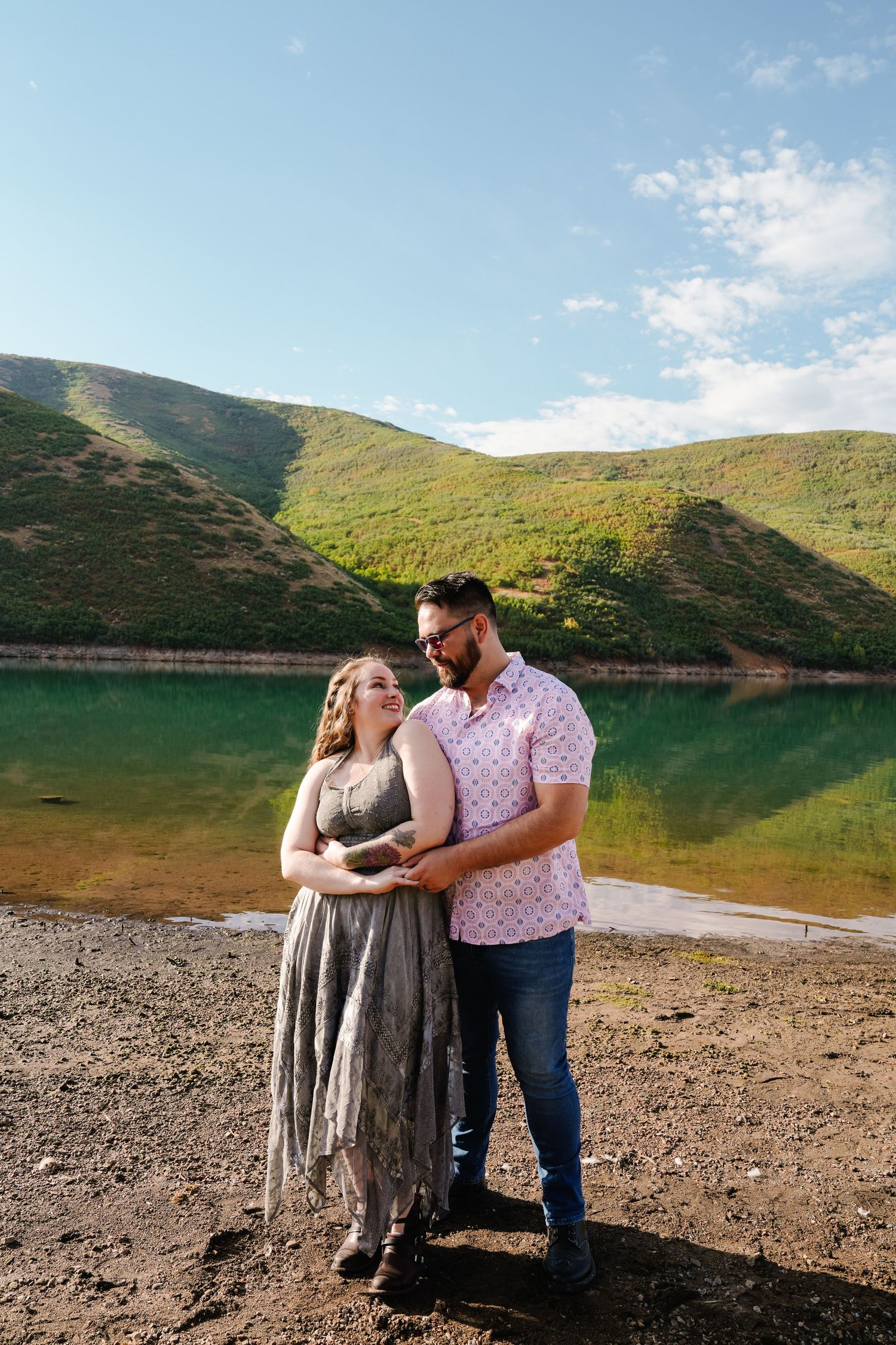 Couple standing together on a beach with green mountains and turquoise lake waters in view.
