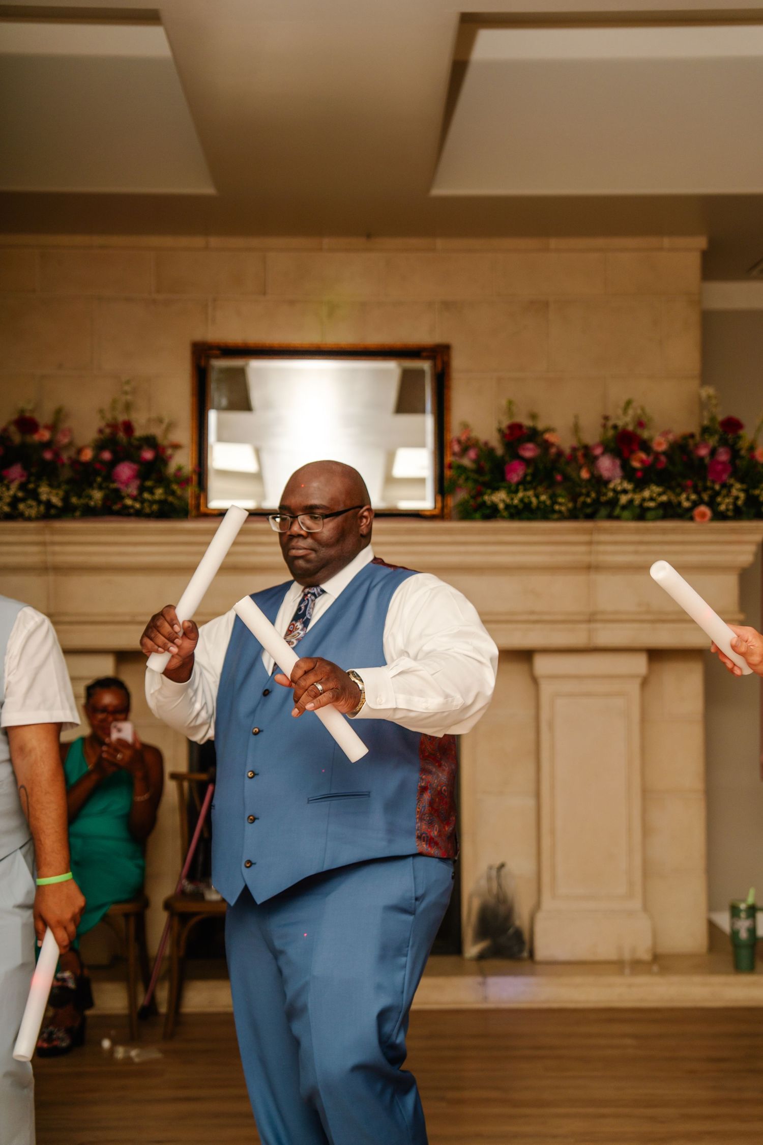 Wedding guest in blue suit vest dancing with light-up wands during reception celebration.