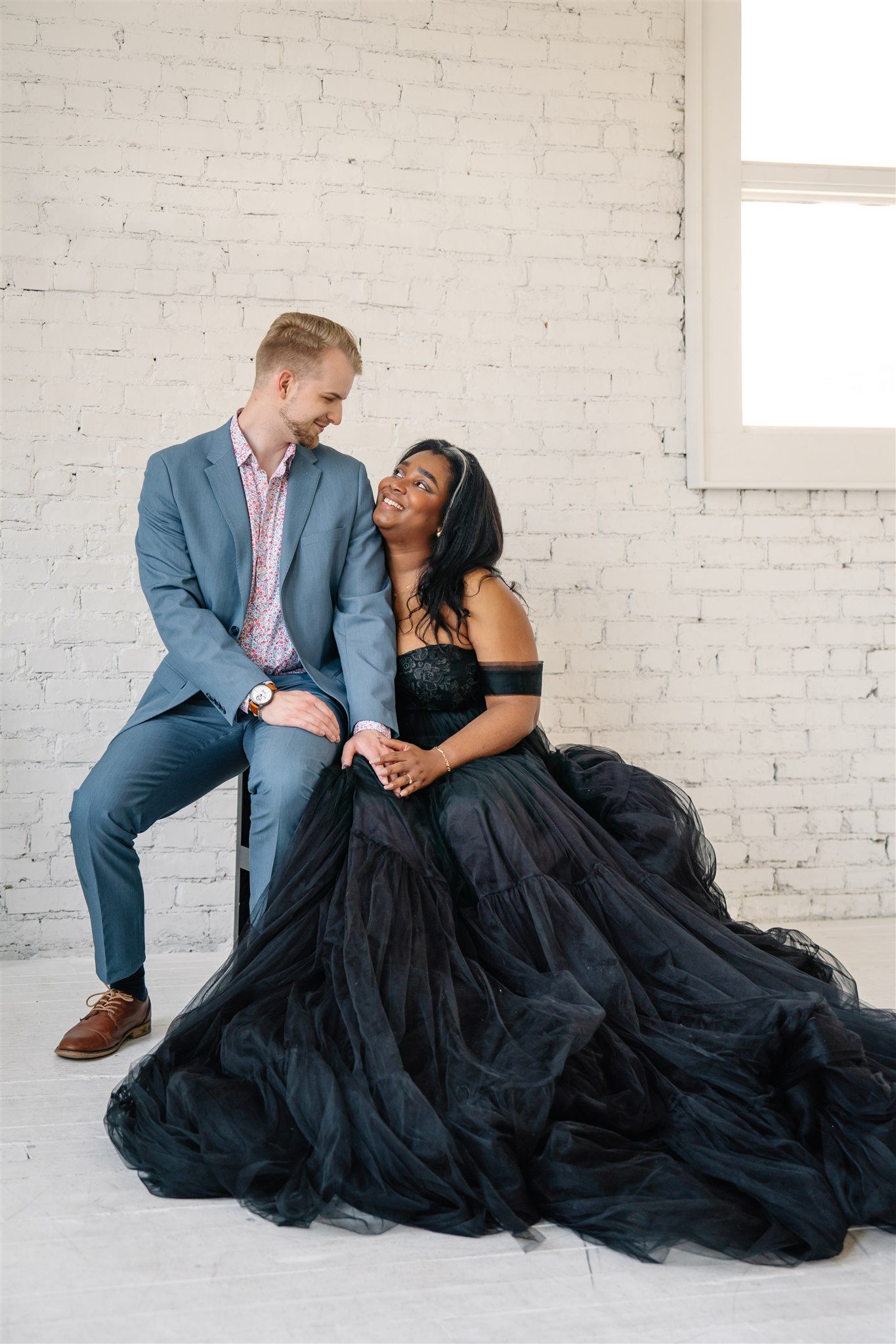 Couple in formal attire posing intimately against white brick wall with flowing black dress.