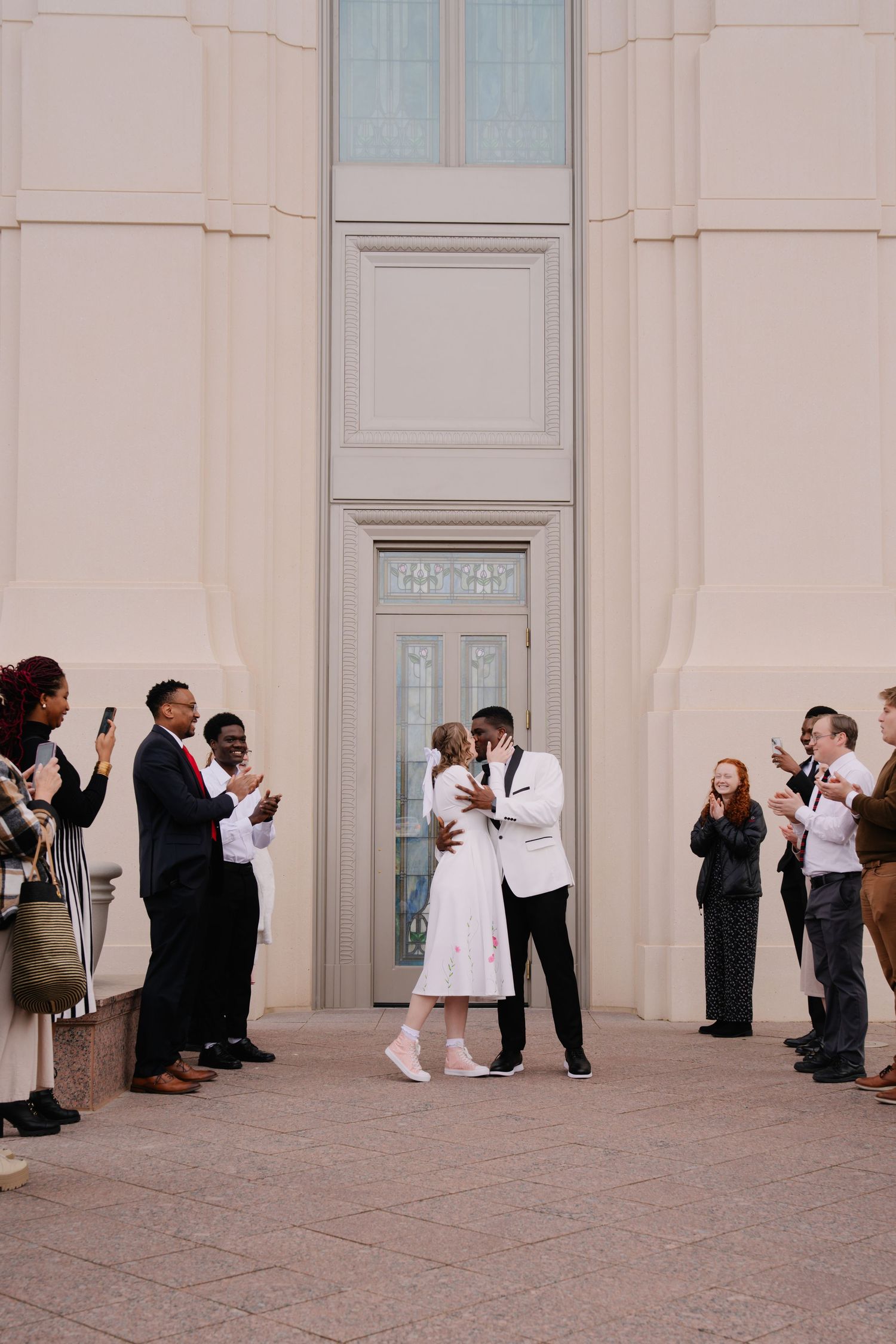Wedding party and guests lined up against white building exterior for photos.