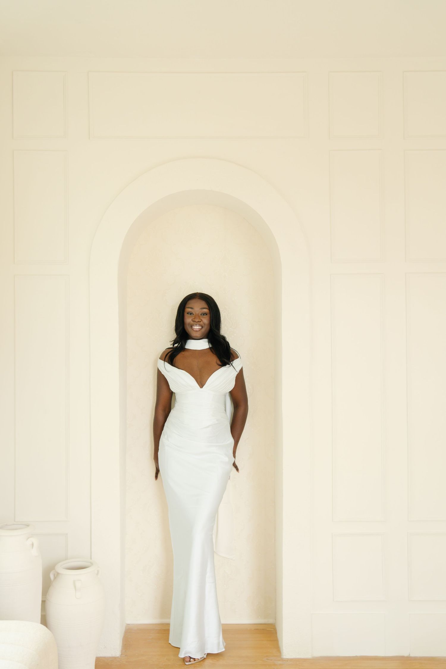 A smiling bride in a white off-shoulder gown stands in an elegant arched alcove against a cream wall.