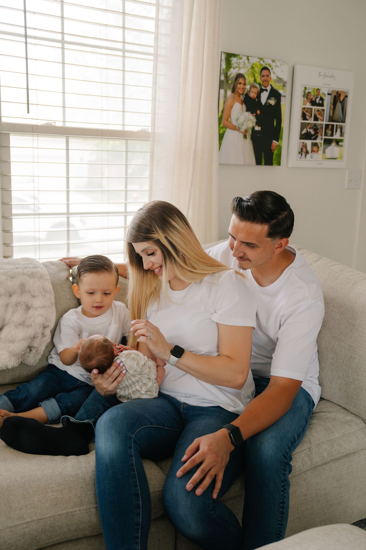 Young family with toddler and newborn baby sitting on couch in bright living room with wedding photo on wall.