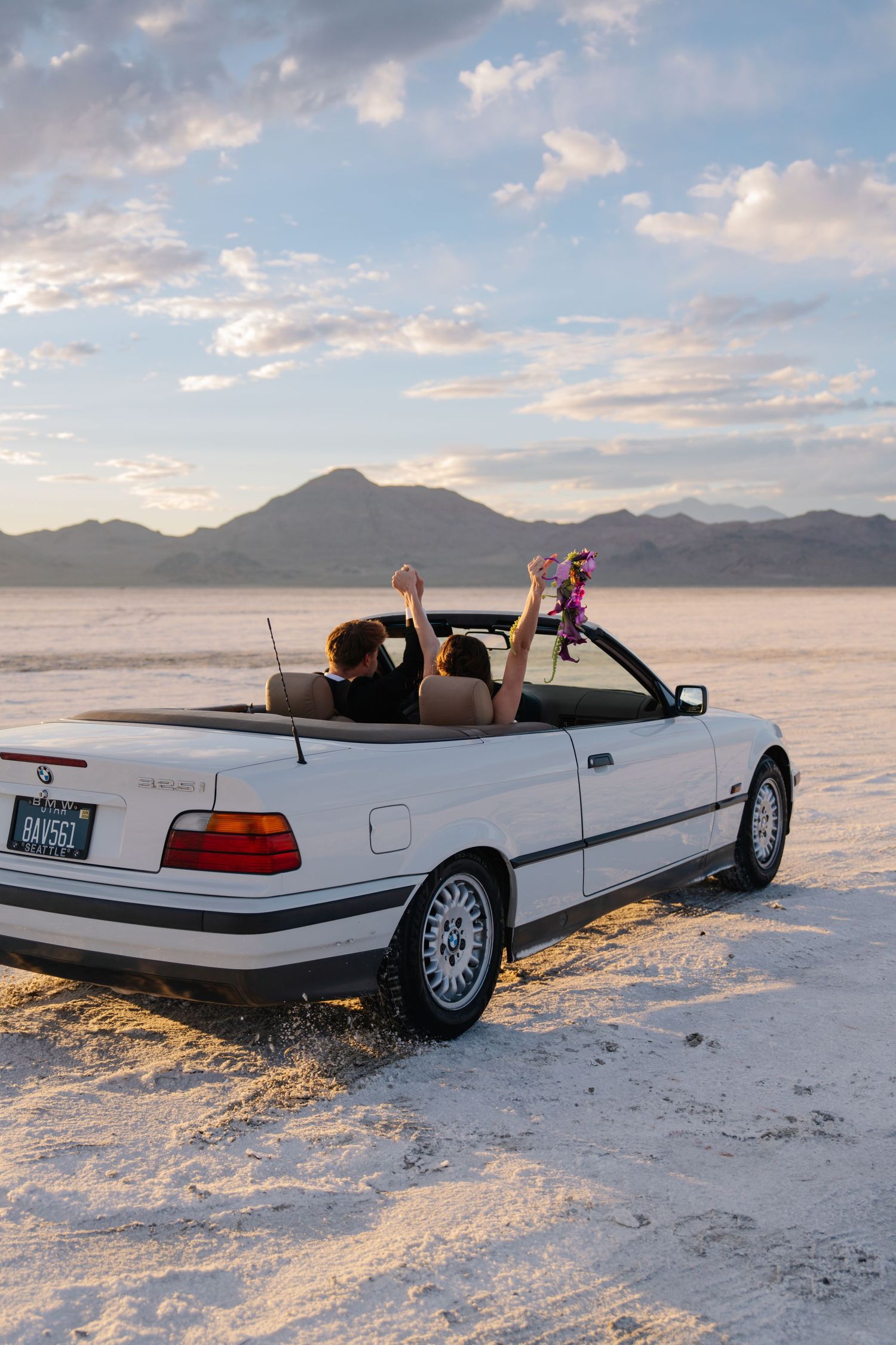 Two people celebrate with raised arms in a white BMW convertible on the Bonneville Salt Flats at sunset.