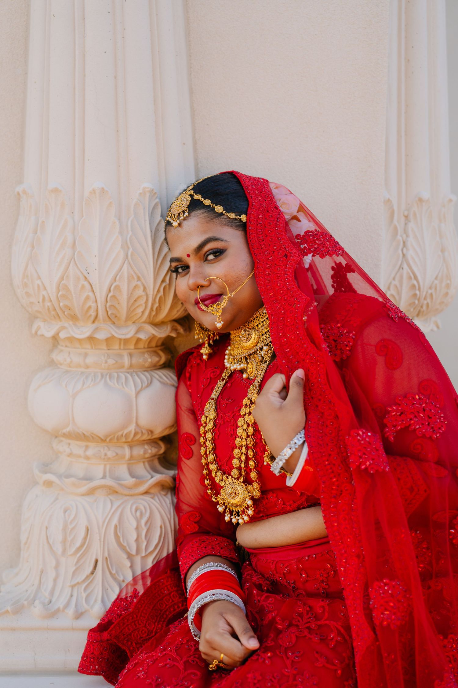 A bride in red traditional attire poses against ornate white marble columns.