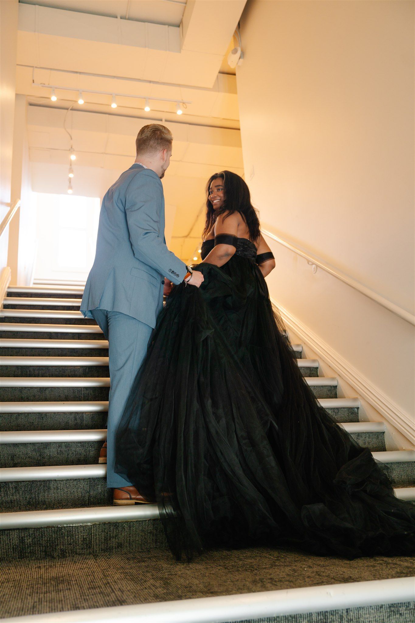 Couple in formal wear posing on illuminated staircase with dramatic black gown.