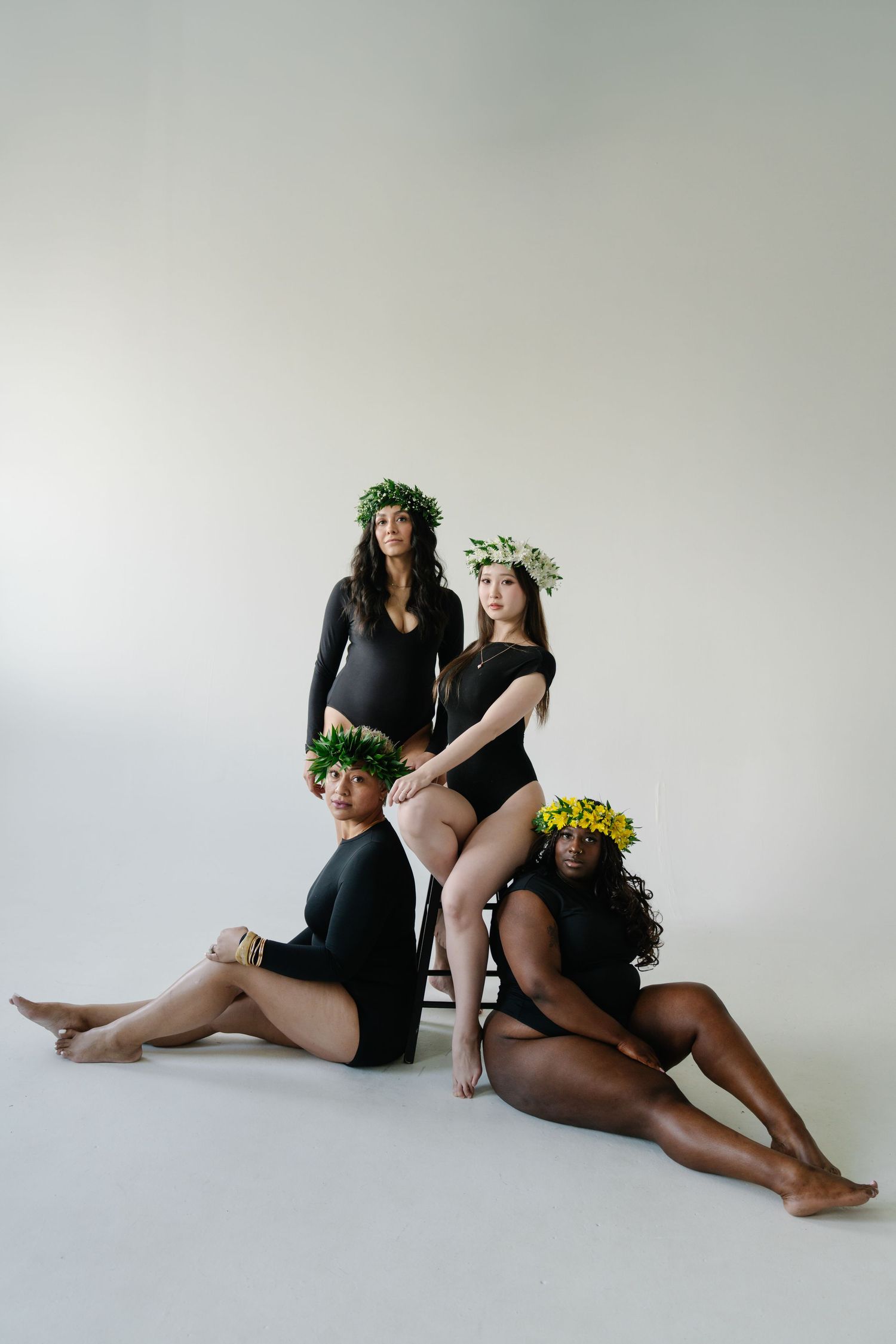 Four diverse women in black bodysuits and floral crowns pose together in a studio setting.
